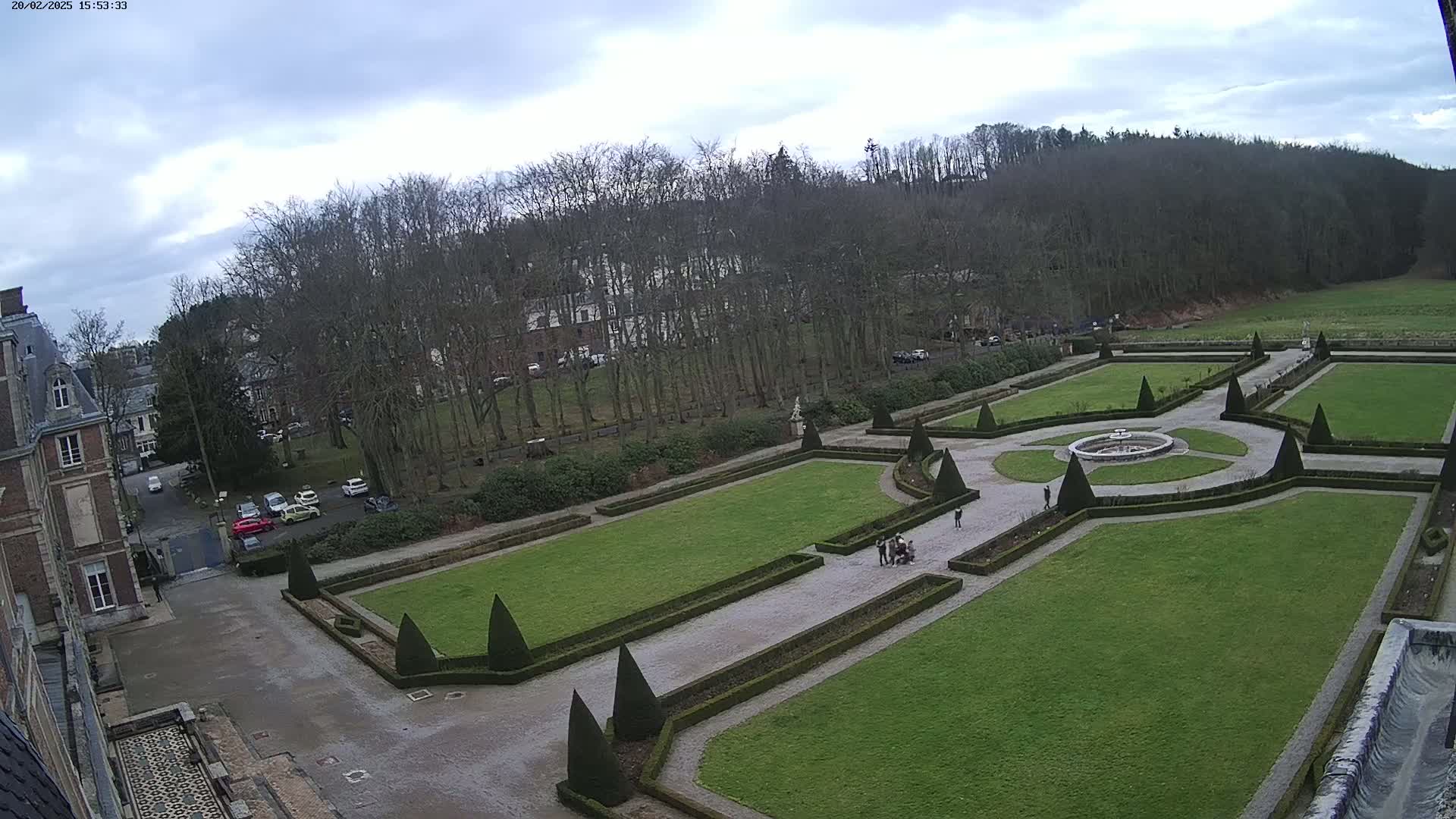 An expansive, formal garden with manicured lawns, geometric hedges, and a central fountain is situated behind a large chateau under a cloudy sky.