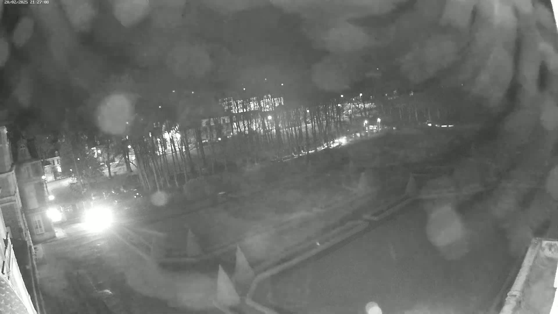 A nighttime, blurry, outdoor security camera view shows a dark roadway, a building, and trees with some lights visible, in snowy or rainy conditions.