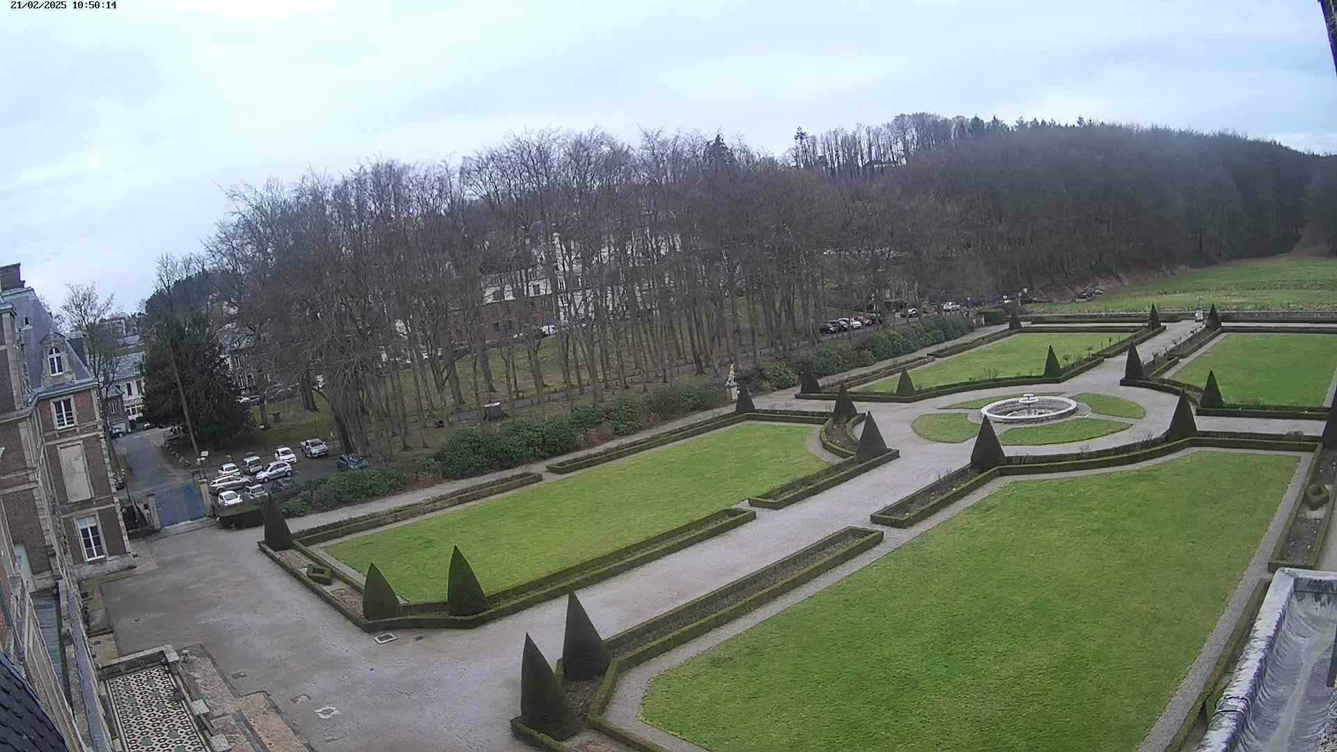 An overcast day reveals a formal garden with geometrically shaped lawns and hedges surrounding a central fountain, adjacent to a large building and a wooded area.