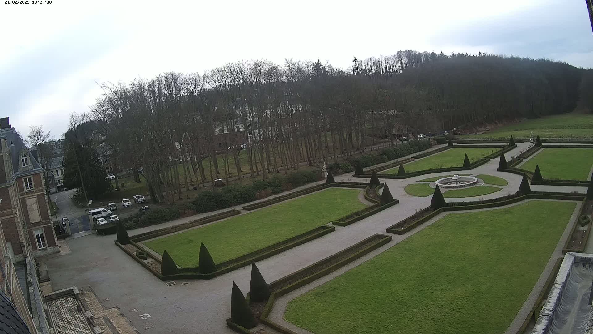 An overcast day reveals an expansive formal garden with geometrically arranged lawns and hedges, centered around a fountain, adjacent to a large building and a line of bare trees.