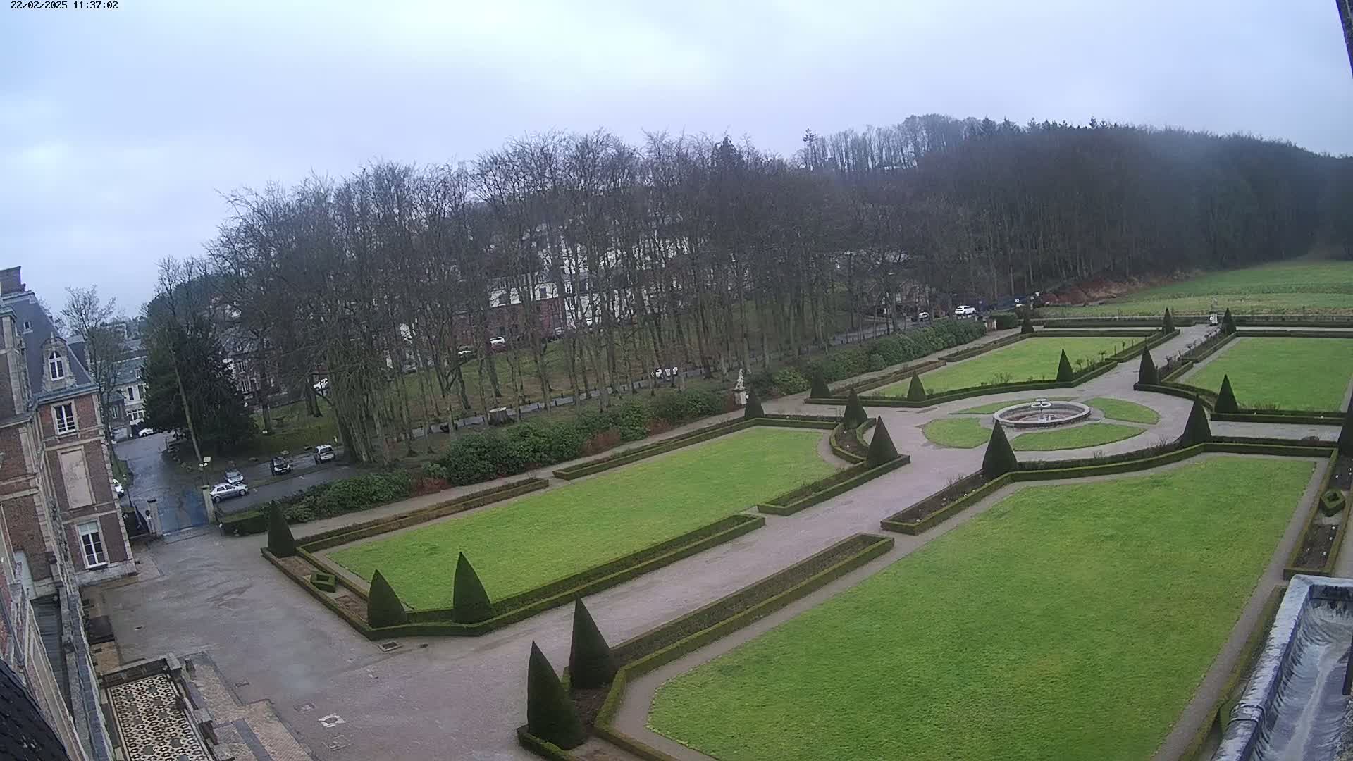 An overcast day reveals a formal garden with geometrically shaped lawns and hedges, adjacent to a large building and a line of trees beyond.