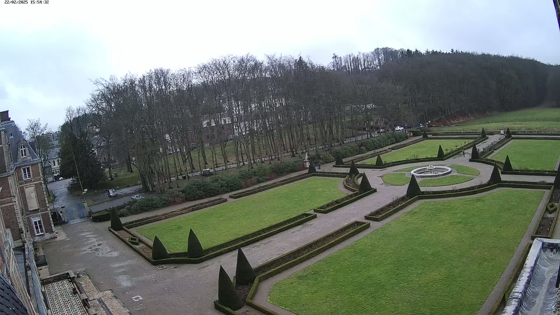 A formal garden with manicured lawns, hedges, and a fountain is situated beside a chateau under an overcast sky.