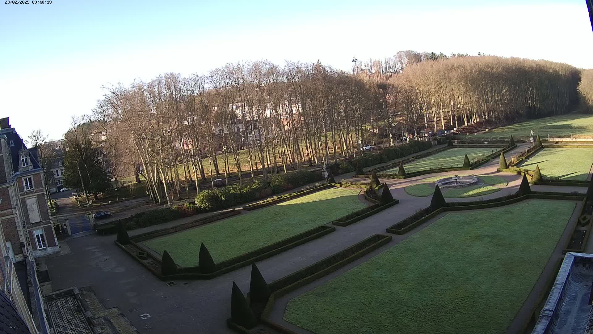 A high-angle view of a formal garden with manicured lawns, hedges, and a fountain, situated next to a large building on a clear, sunny day.