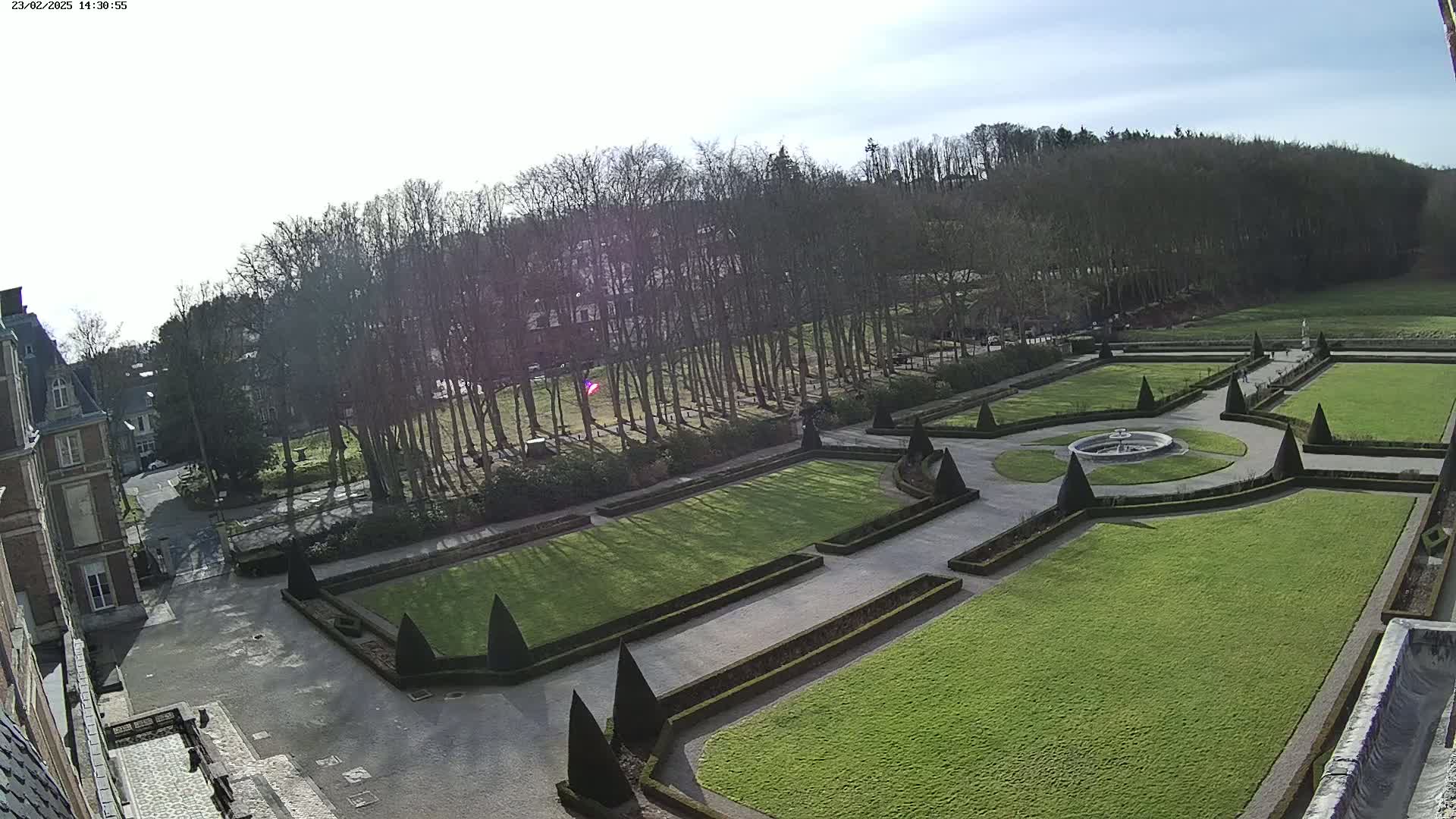 A formal garden with manicured lawns, geometric hedges, and a central fountain is visible from an elevated viewpoint on a sunny day, with a large building and trees in the background.