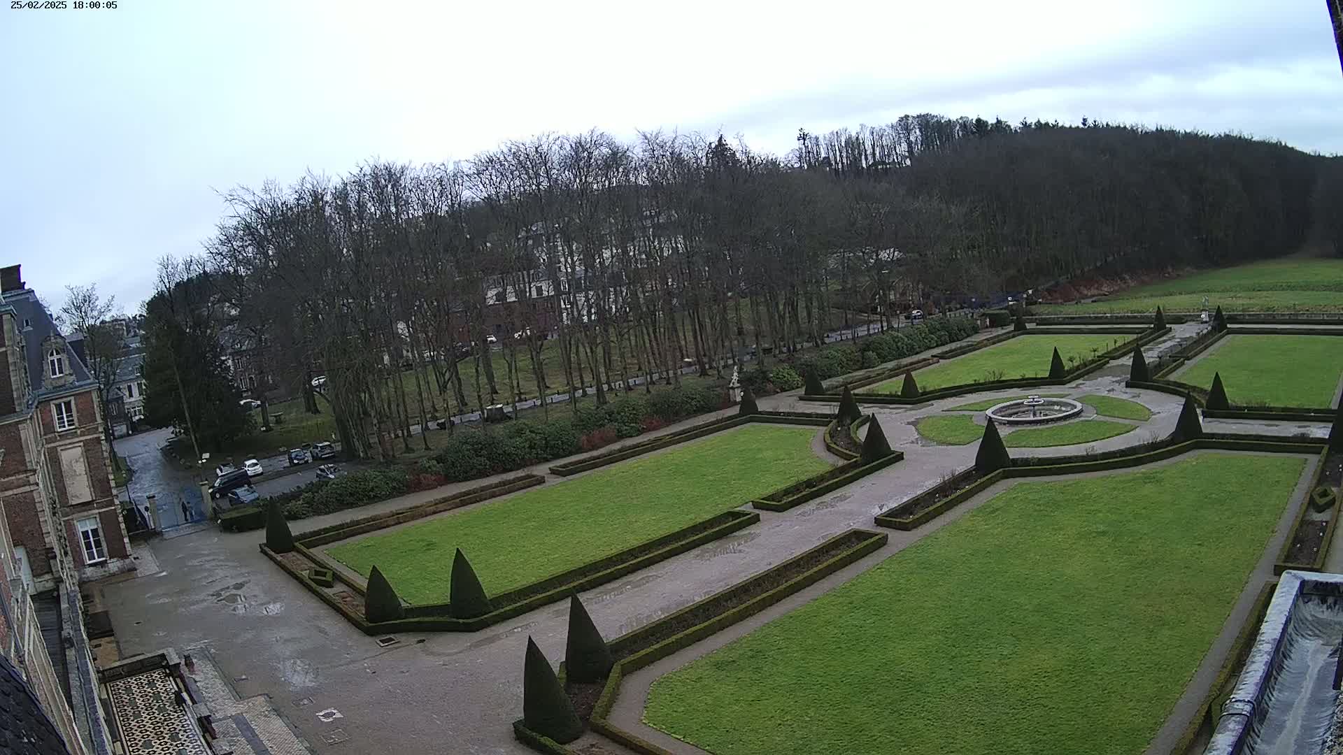 An expansive formal garden with manicured lawns, hedges, and a central fountain is situated beside a large building under a cloudy sky.