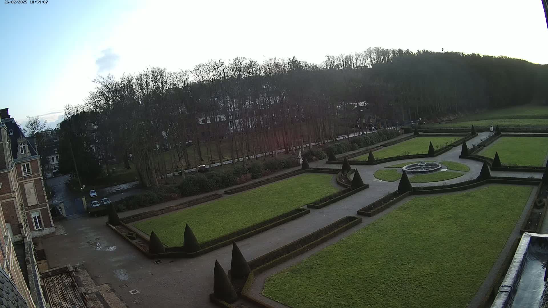 An aerial view shows a formal garden with manicured lawns, hedges, and a fountain, situated next to a large building and a line of bare trees under a mostly clear sky.