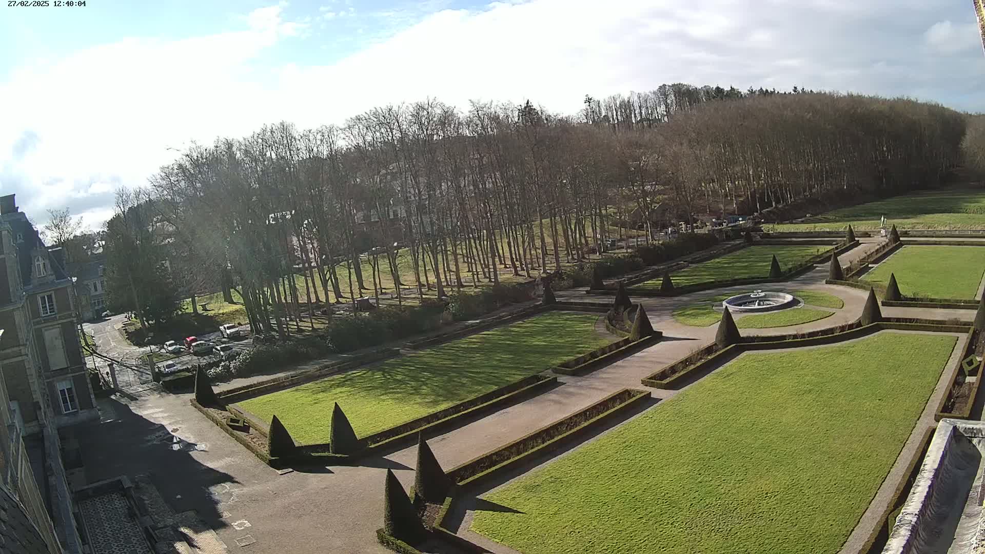 An aerial view of a formal garden with manicured lawns, hedges, and a central fountain, situated next to a building and bordered by a line of leafless trees under a partly sunny sky.