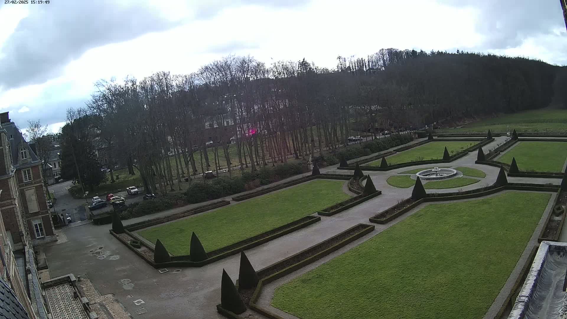 An aerial view of a formal garden with a central fountain, bordered by manicured lawns, hedges, and pathways, situated beside a building under a cloudy sky.