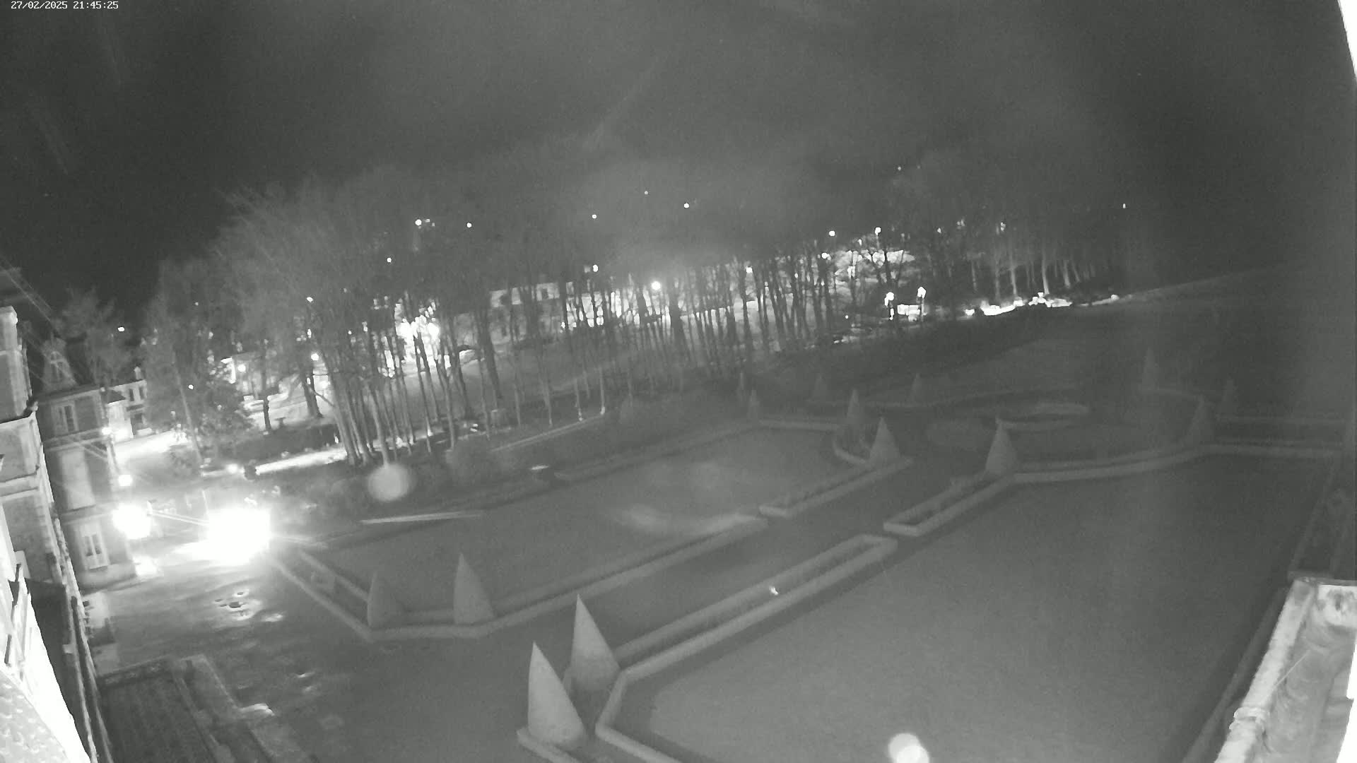A nighttime, low-light view of a geometrically patterned garden, bordered by trees and buildings, under a dark sky.