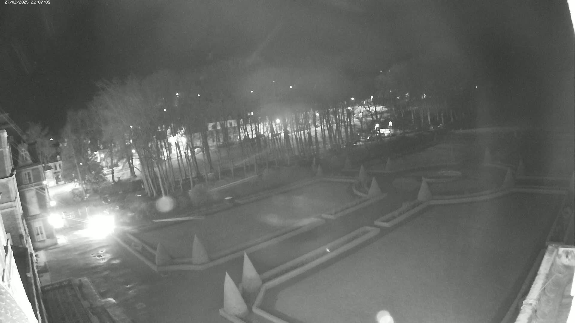 A nighttime, low-light view of a formal garden with geometric hedges and a building in the background, possibly under a clear sky.