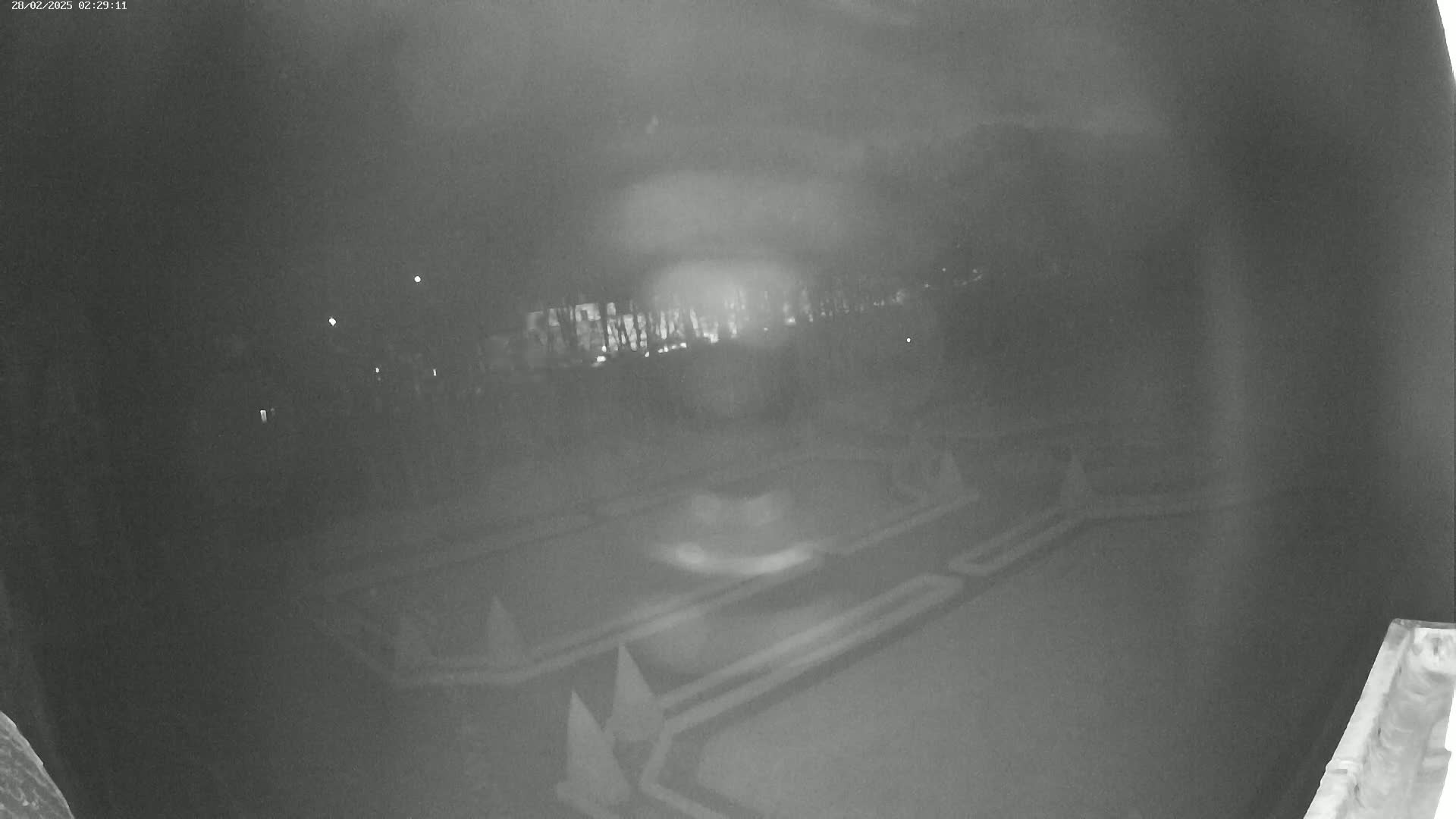 A nighttime, low-light security camera view shows an empty outdoor mini golf course with blurry lights in the background.
