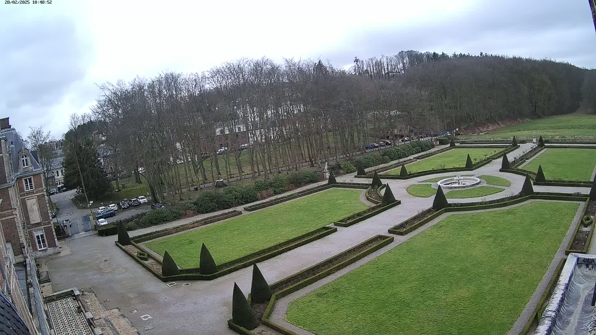 A formal garden with a central fountain is situated next to a large building under an overcast sky.
