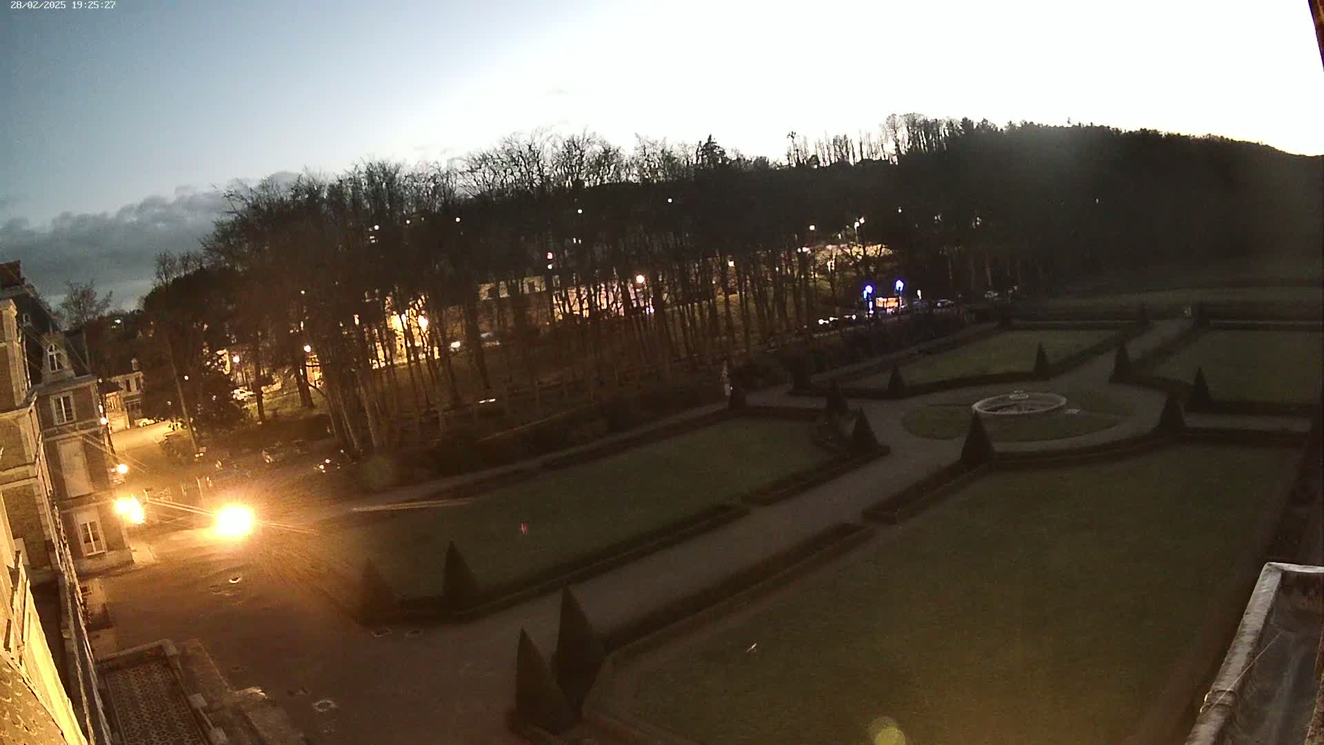 A nighttime view of a formal garden with geometrically shaped lawns and hedges, situated near a building and a line of trees under a twilight sky.