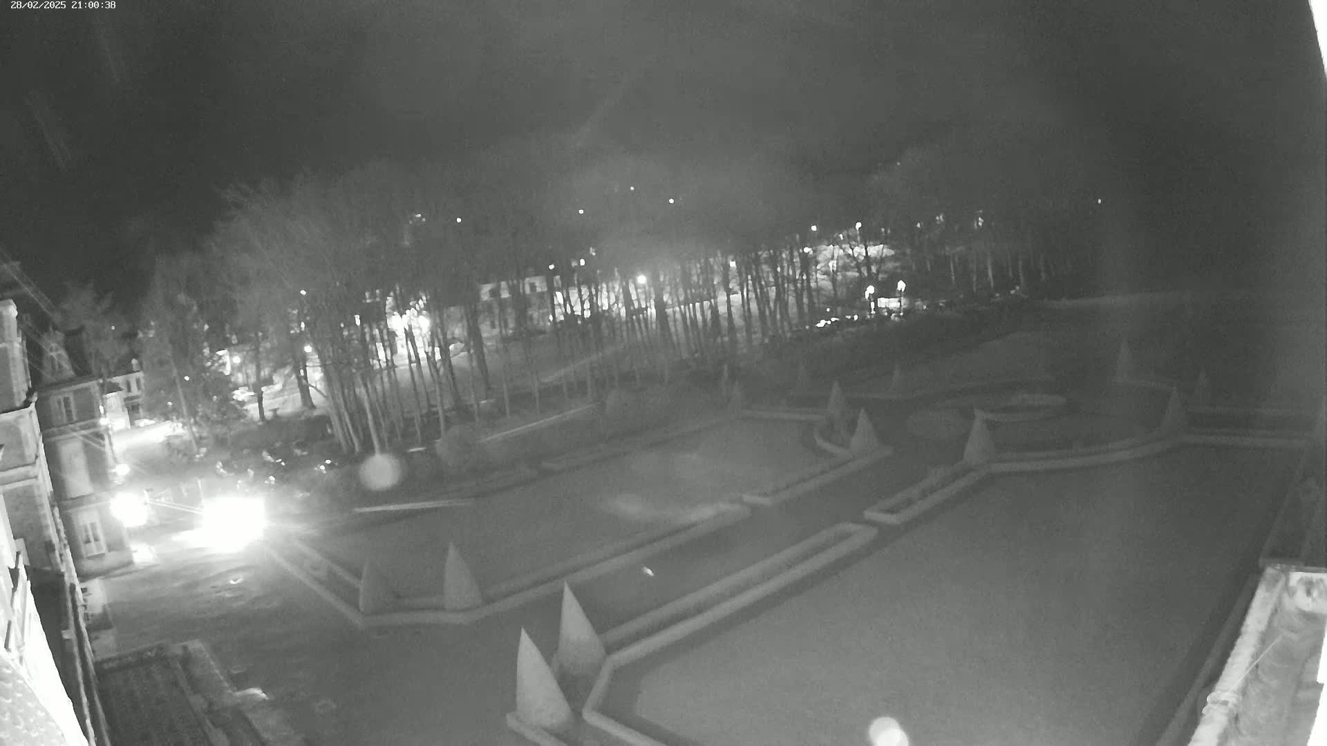 A nighttime, black and white overhead view shows a formal garden with geometric patterns, surrounded by trees and buildings under a dark sky.