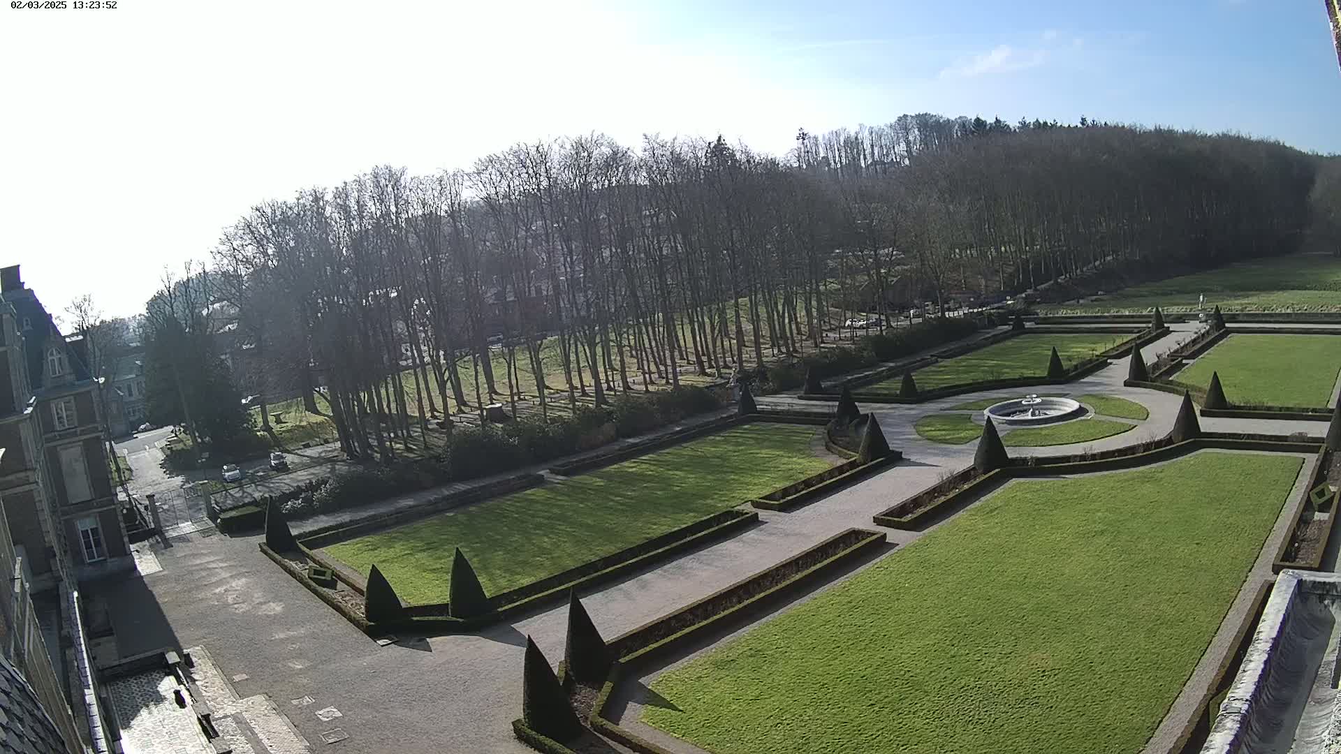 A formal garden with manicured lawns, hedges, and a fountain is situated in front of a chateau under a sunny sky.