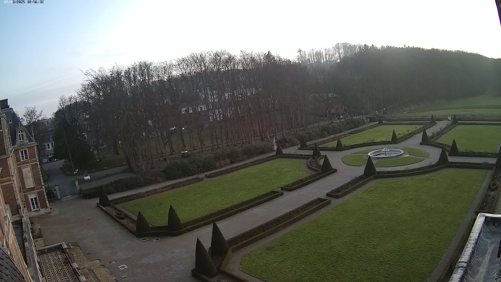 An ornate formal garden with a central fountain is situated beside a large brick building on an overcast day.