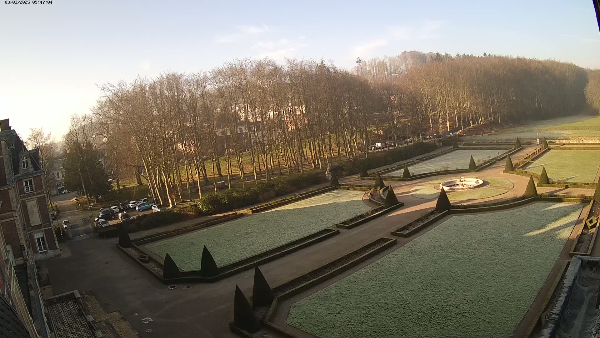 A frost-covered formal garden with a central fountain is situated in front of a chateau,  backed by a line of leafless trees and a hillside under a clear, bright sky.
