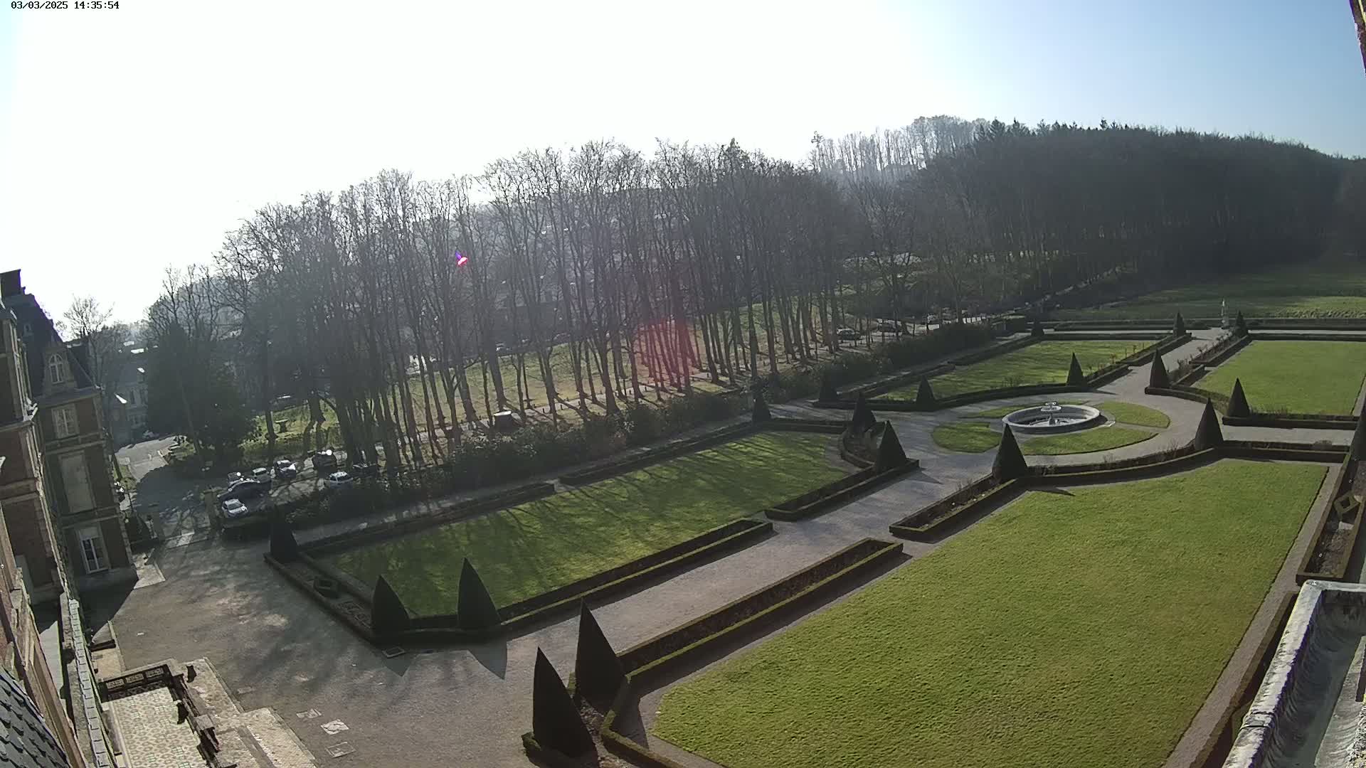 A formal garden with a central fountain is visible from an elevated viewpoint on a sunny day, bordered by a large building and a line of leafless trees.