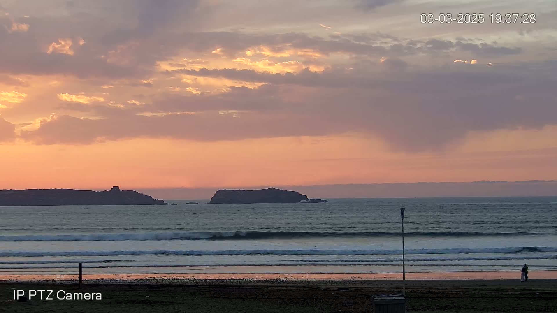 A pink and orange sunset over a calm ocean with small waves lapping a sandy beach, two figures standing near the water's edge, and a small island in the distance.