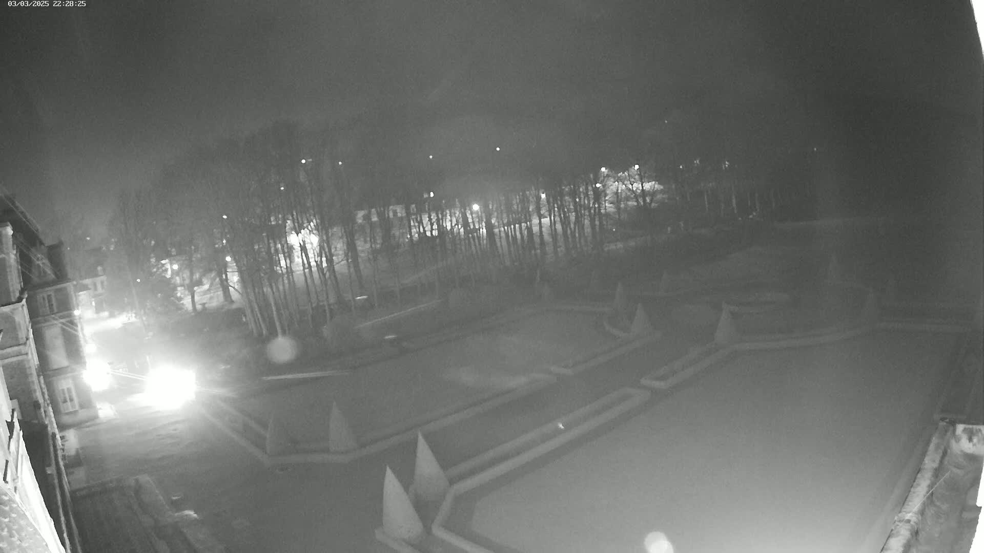 A nighttime, low-light view shows a formal garden with geometrically shaped beds, bordered by trees and a building, illuminated by faint lights.