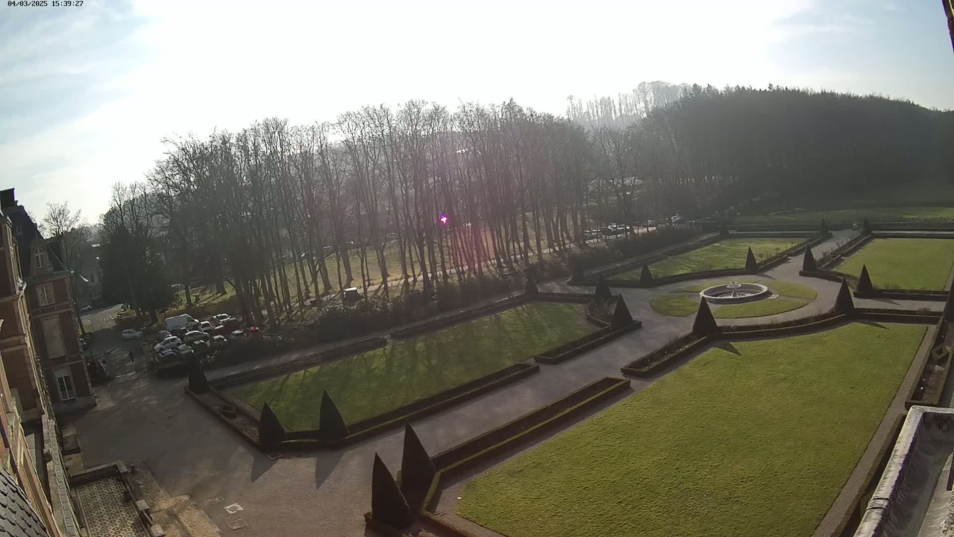 A high-angle view of a formal garden with a fountain, manicured lawns, and hedges, situated beside a building under a bright, sunny sky.