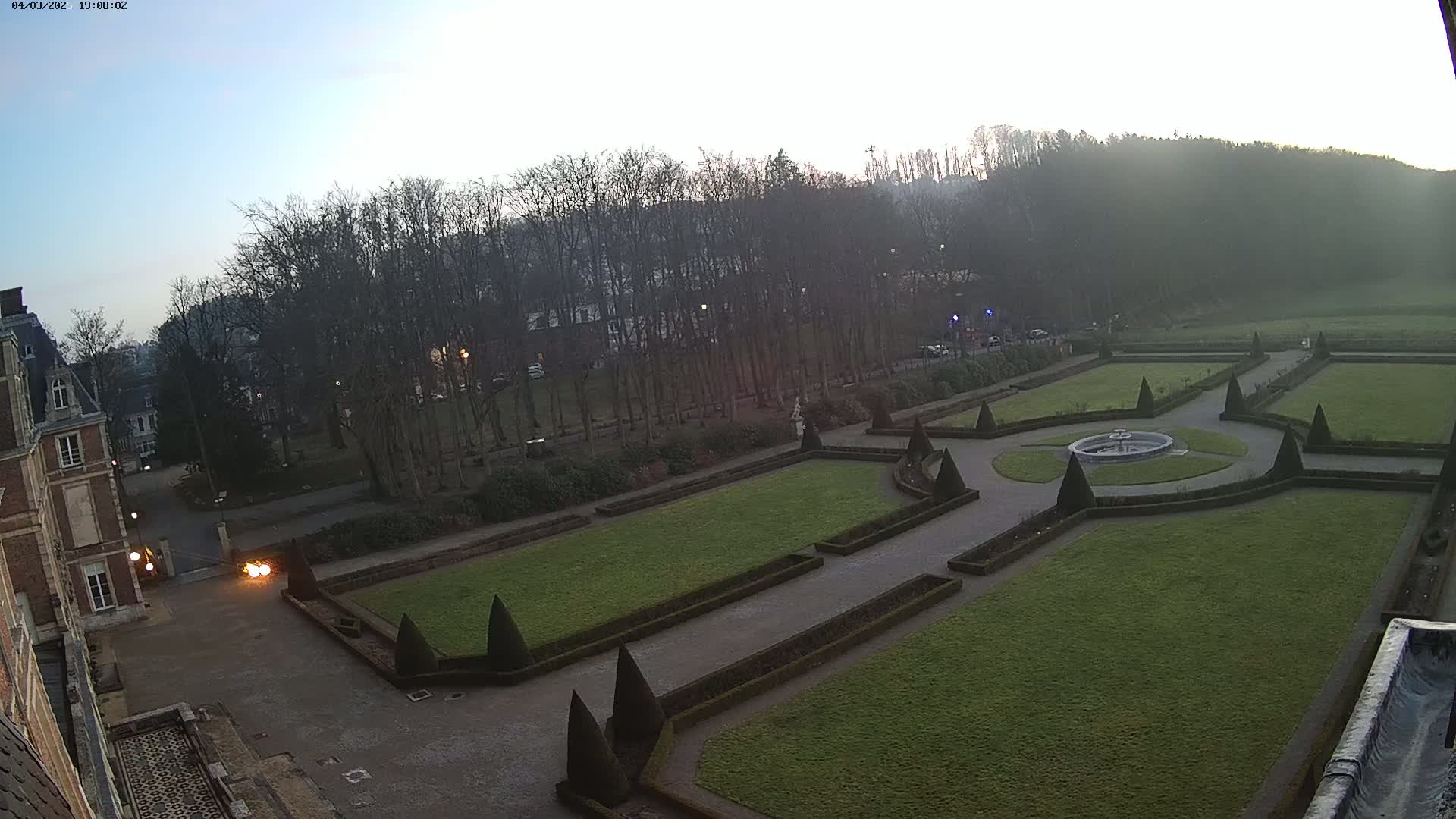 A formal garden with a central fountain, manicured lawns, and hedges is situated next to a large brick building under a clear, dusky sky.