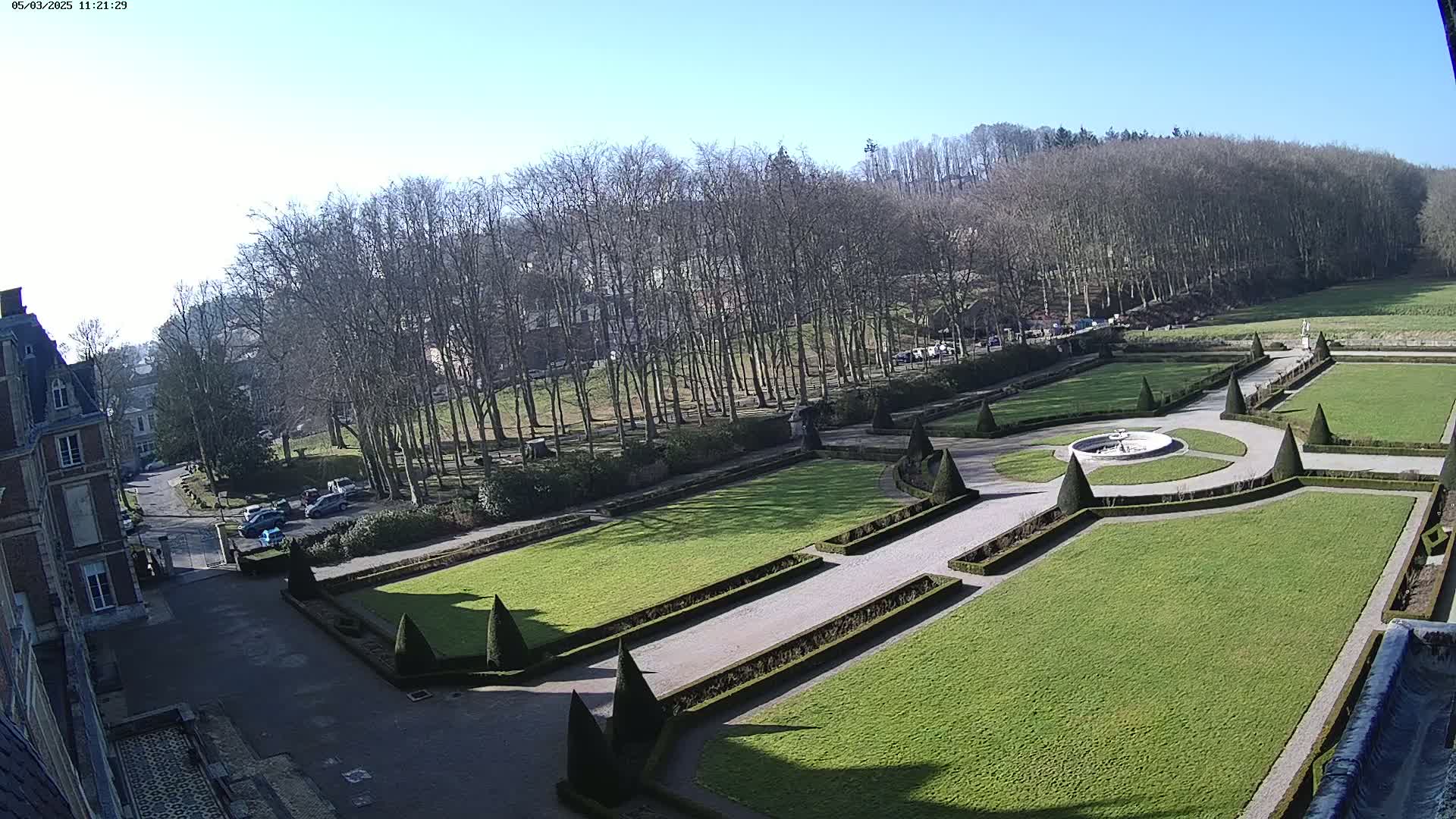 A formal garden with manicured lawns, hedges, and a fountain is situated below a chateau on a sunny day.