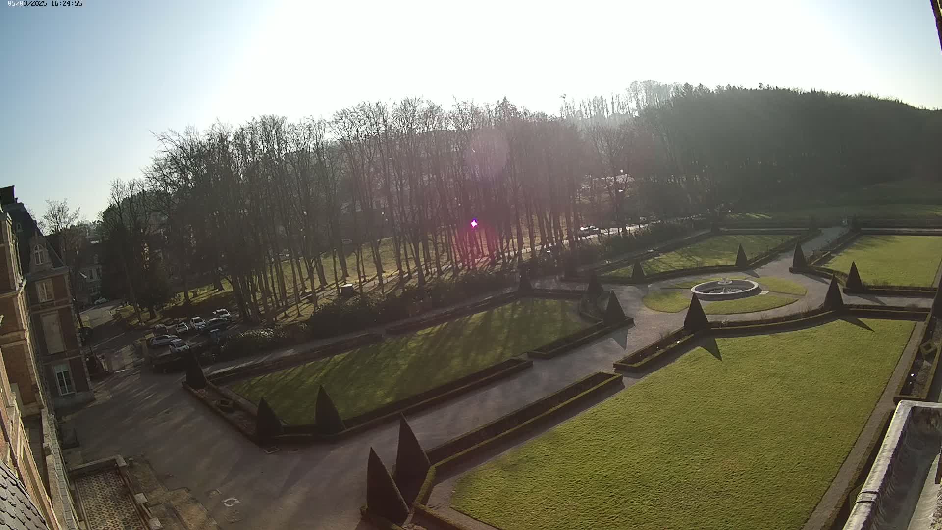 A sunny day reveals a formal garden with manicured lawns, hedges, and a central fountain, adjacent to a building and a stand of leafless trees.
