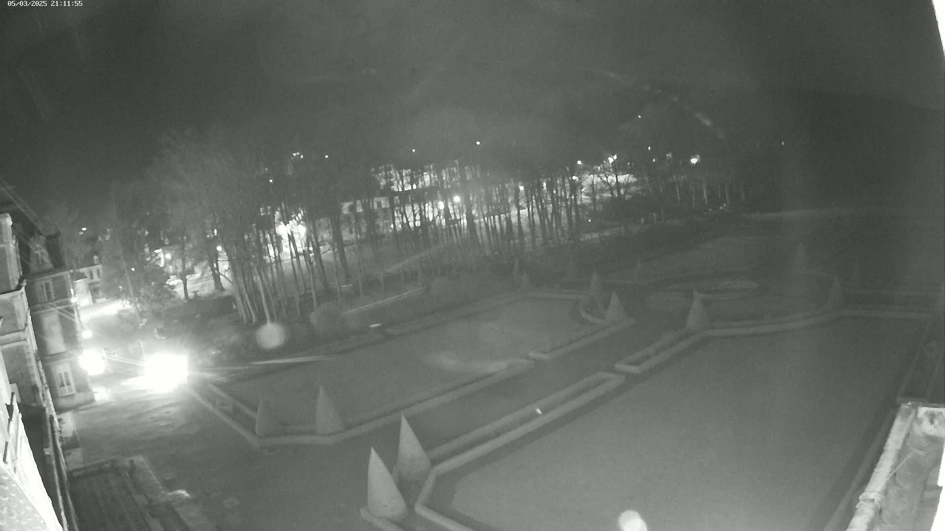 A nighttime, black-and-white view shows a paved area with a geometrically designed garden, trees, and buildings in the background.