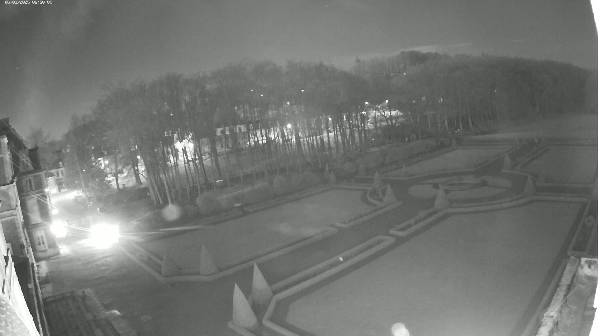 A nighttime, high-angle view shows a formal garden with geometrically-designed hedges and a building in the background under a dark sky.