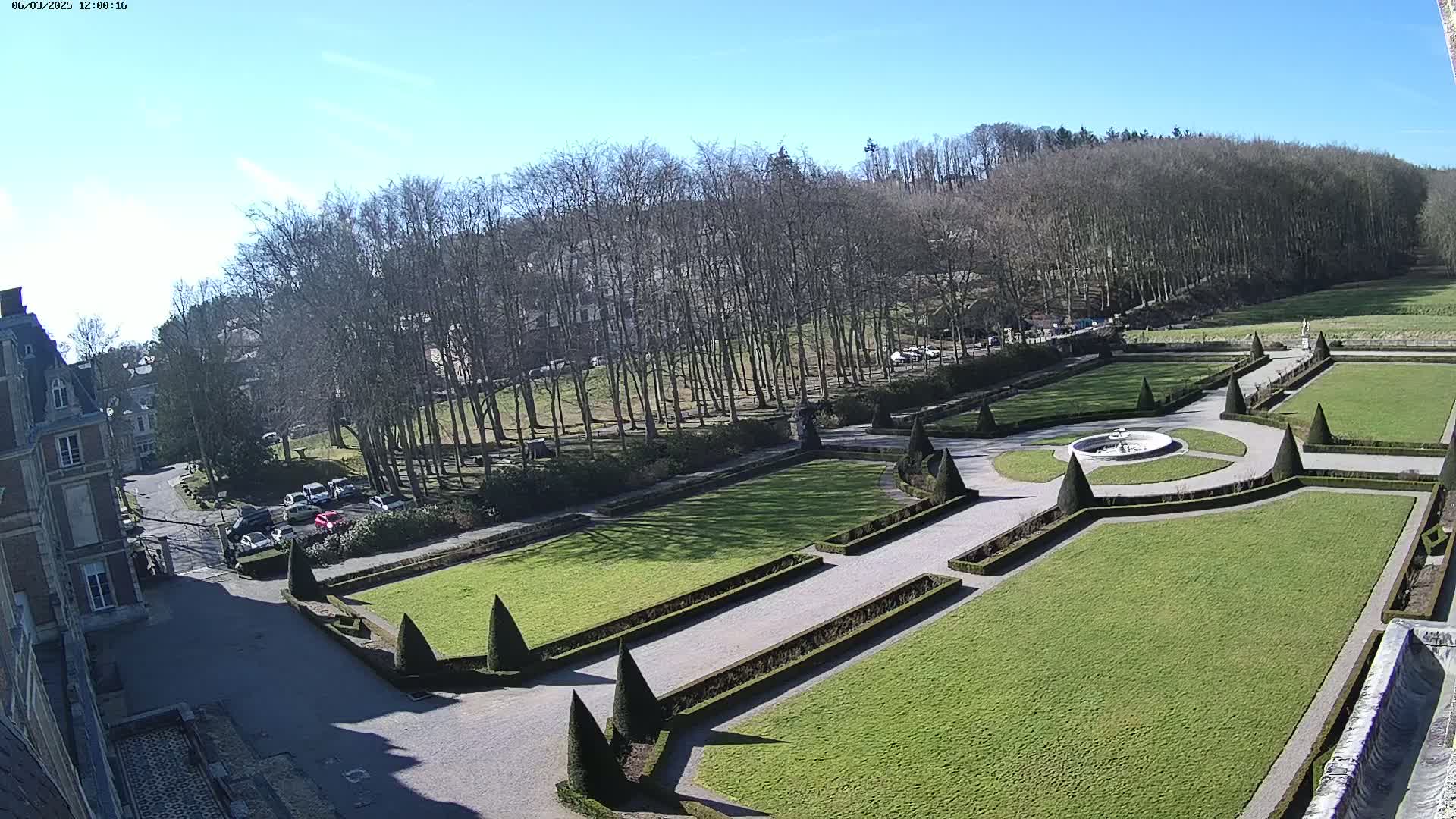 A formal garden with manicured lawns, hedges, and a fountain is situated beside a large building, all under a clear, sunny sky.