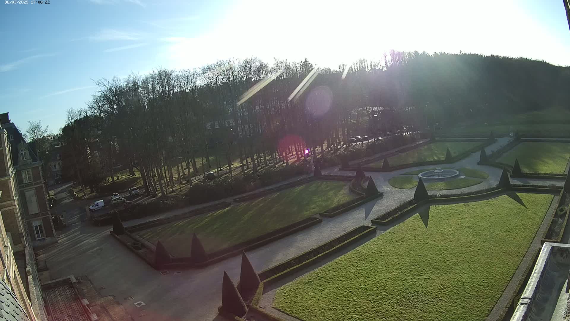 An elevated view of a formal garden with manicured lawns, hedges, and a central fountain, situated beside a large building under a bright, sunny sky.