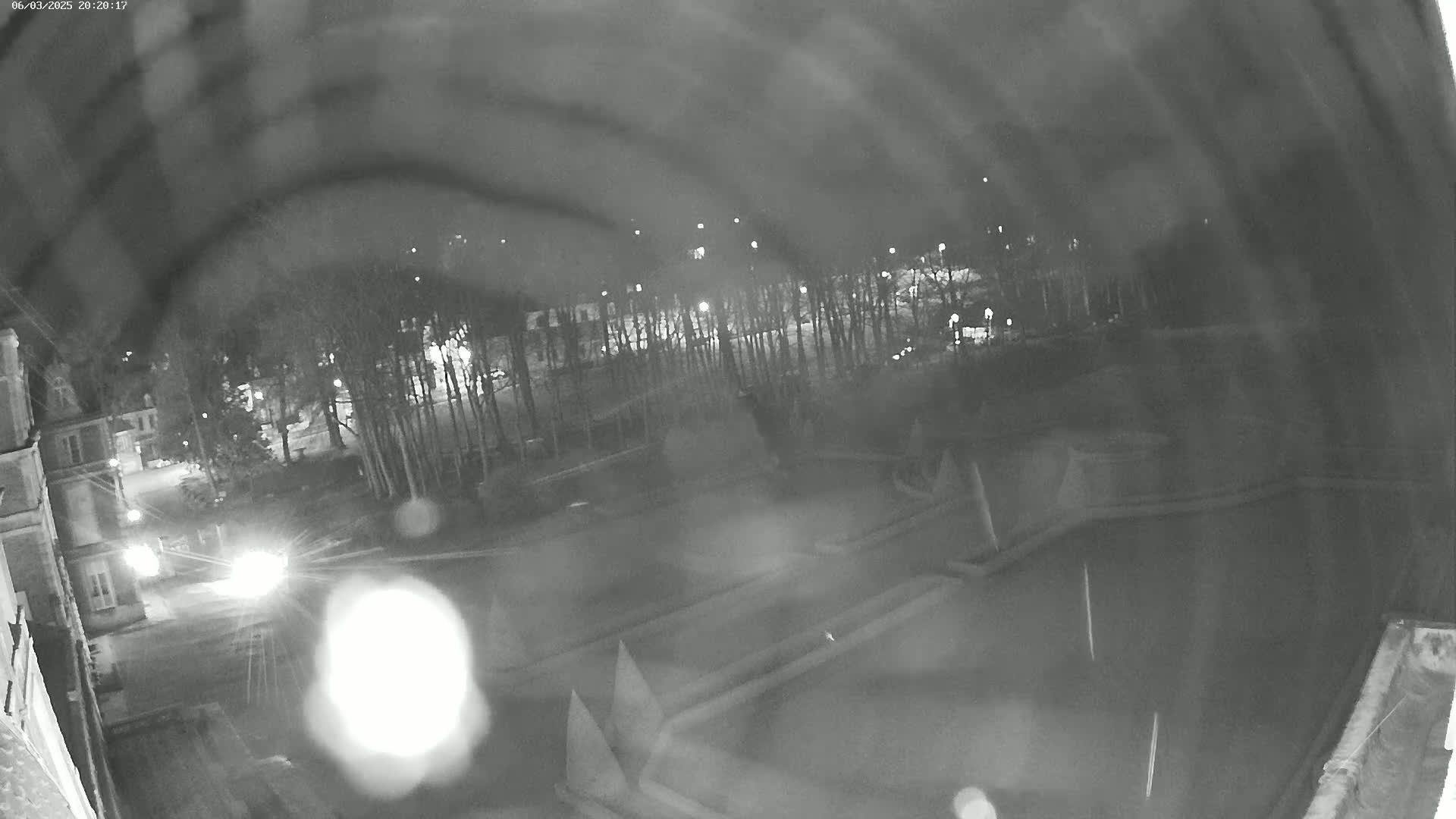 A nighttime, blurry view from a high vantage point shows a dark roadway, buildings, trees, and what appears to be a formal garden or park with geometric shapes, all under dark, possibly cloudy skies.
