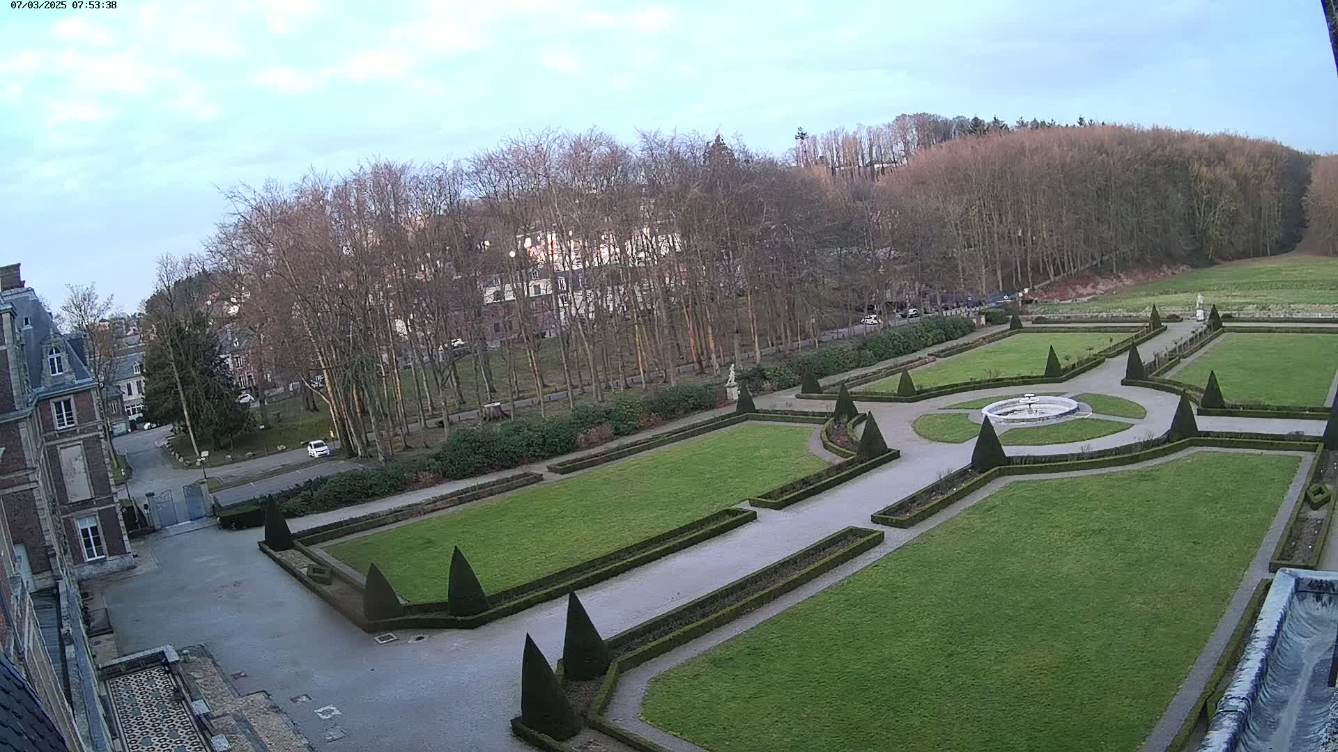 An aerial view of a formal garden with a central fountain, geometrically shaped hedges, and a backdrop of bare trees under a partly cloudy sky.