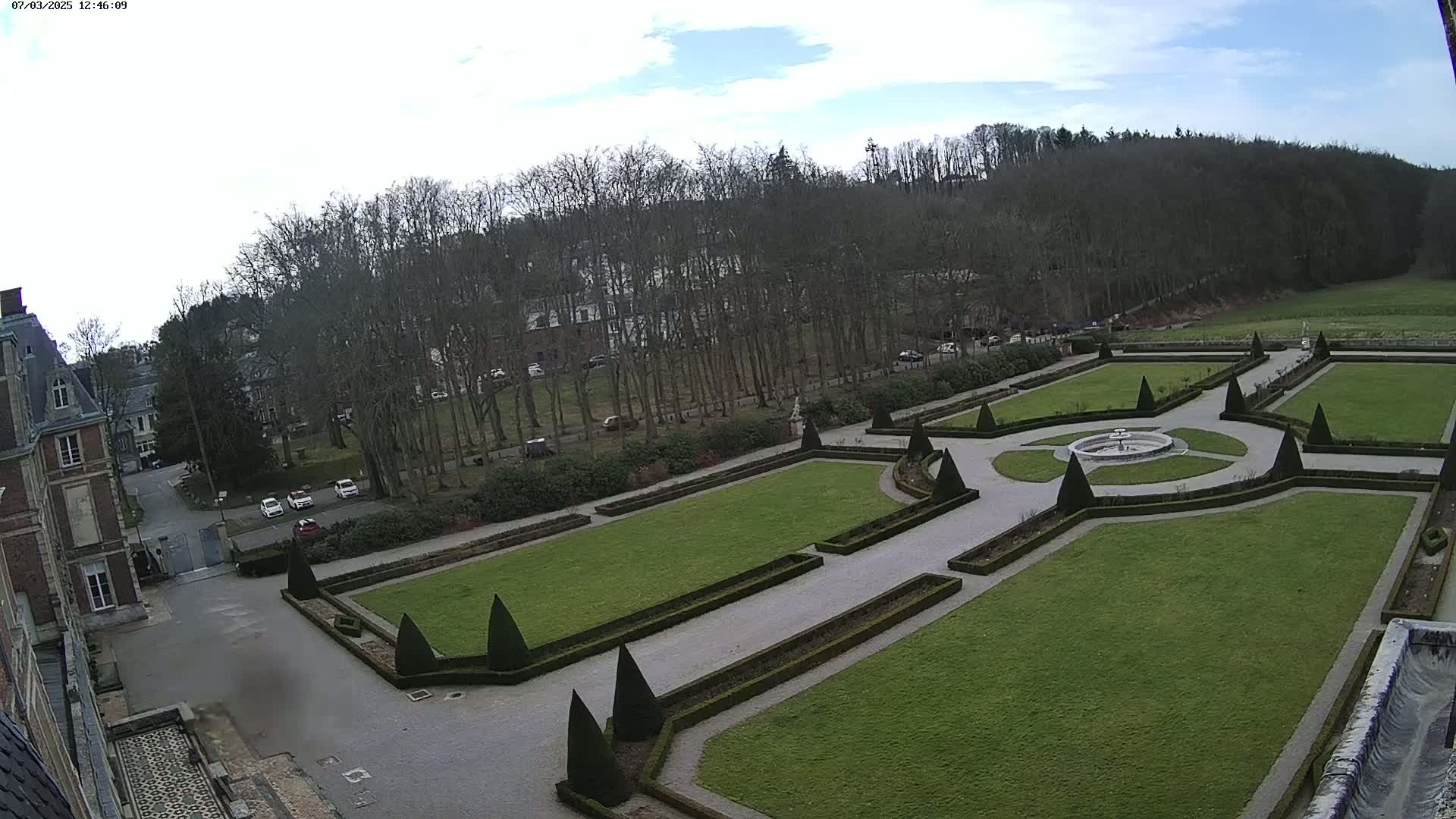 A formal garden with a central fountain is visible from an elevated vantage point,  bordered by a large building and a background of leafless trees under a partly cloudy sky.