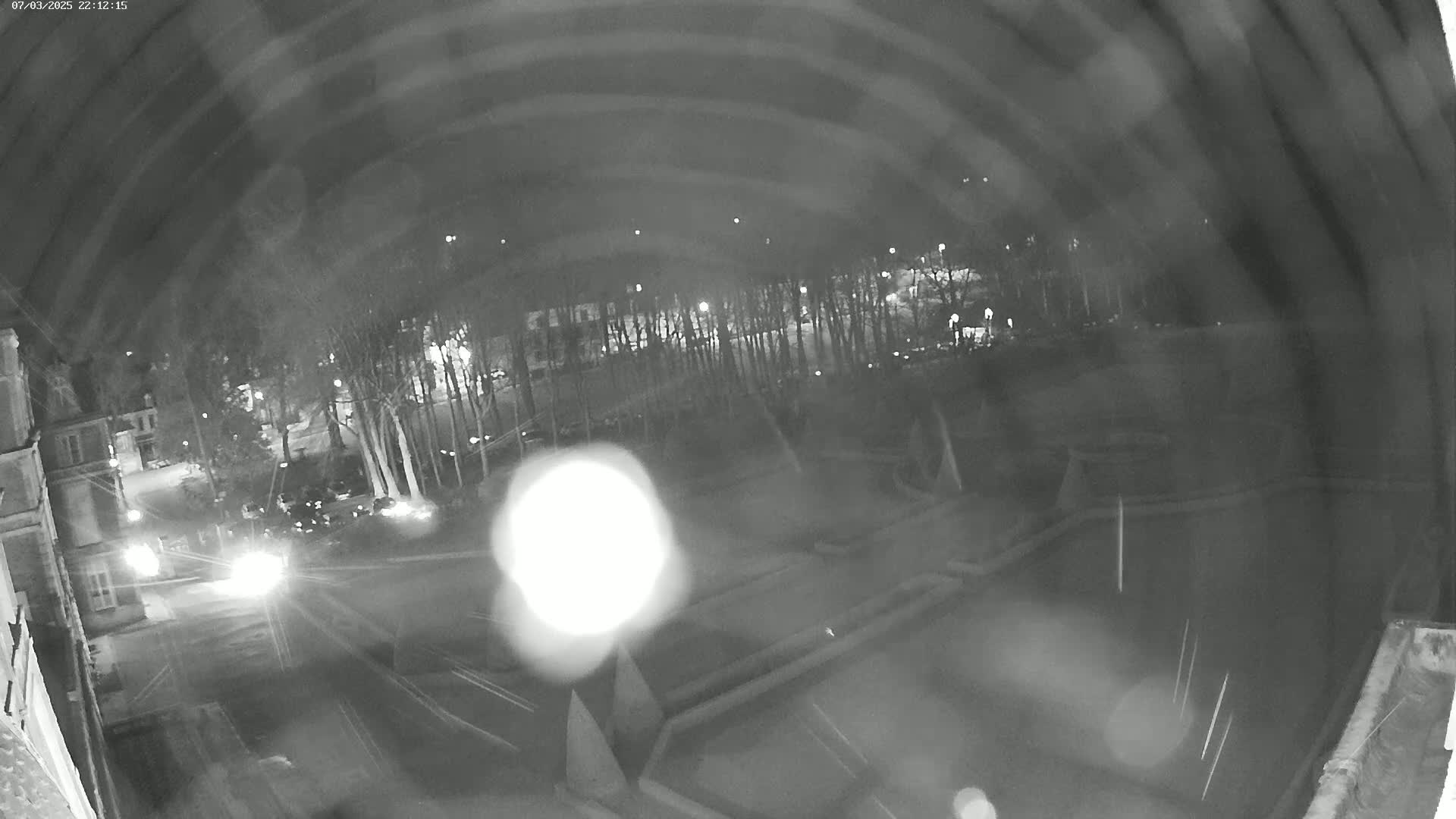 A nighttime, blurry view of a street and park, showing vehicles with headlights on and trees under dim lighting conditions.