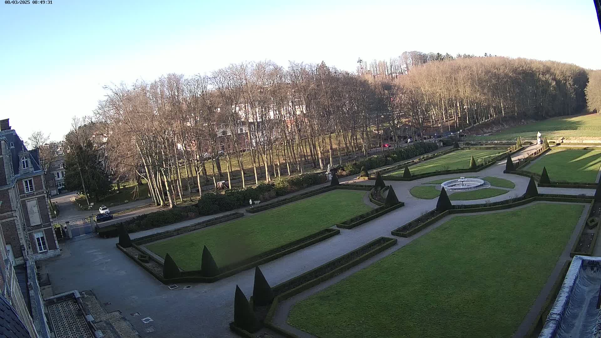A formal garden with manicured lawns and hedges, a fountain, and a building in the background, on a sunny day.