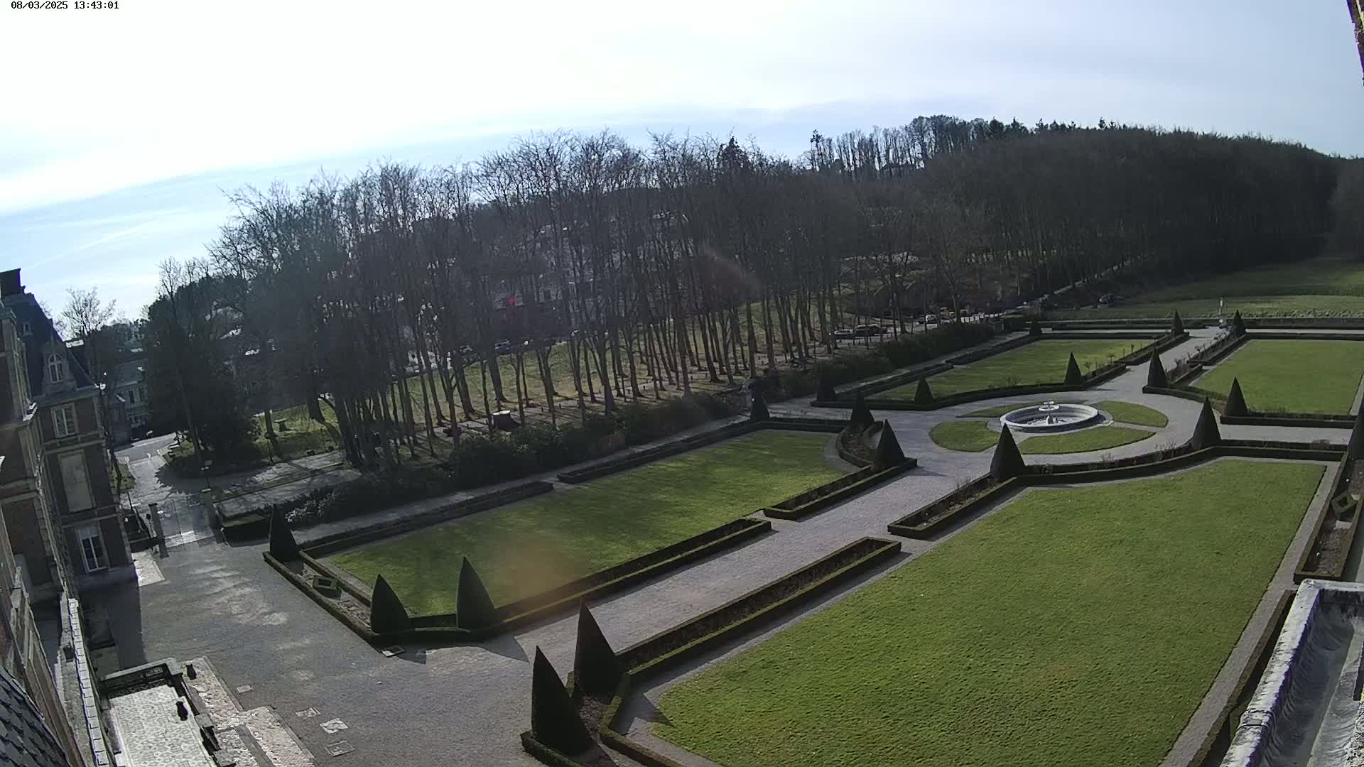 An aerial view shows a formal garden with a central fountain, manicured lawns, and hedges, situated next to a large building under a mostly sunny sky.