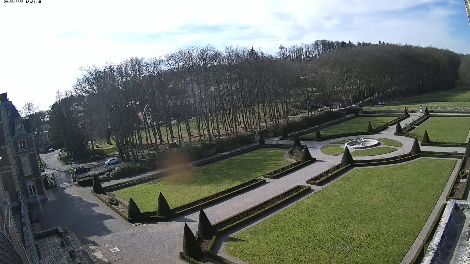 An elevated view shows a formal garden with manicured lawns, hedges, and a central fountain, adjacent to a large building under a mostly sunny sky.