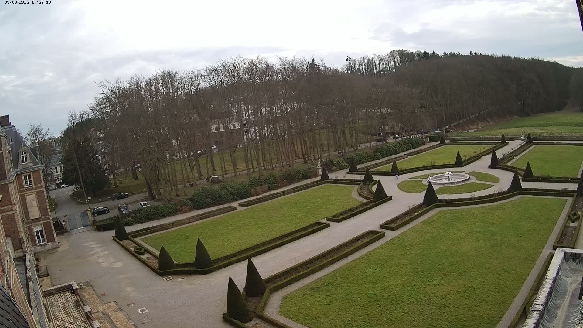 An expansive formal garden with manicured lawns and hedges, situated adjacent to a chateau under an overcast sky.