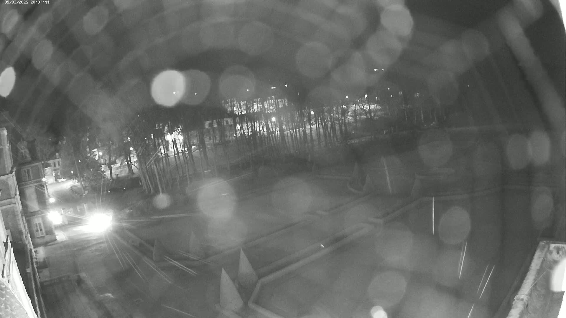 A nighttime, rain-blurred view shows a courtyard with geometric patterns, trees, a building, and a vehicle's headlights in the distance.