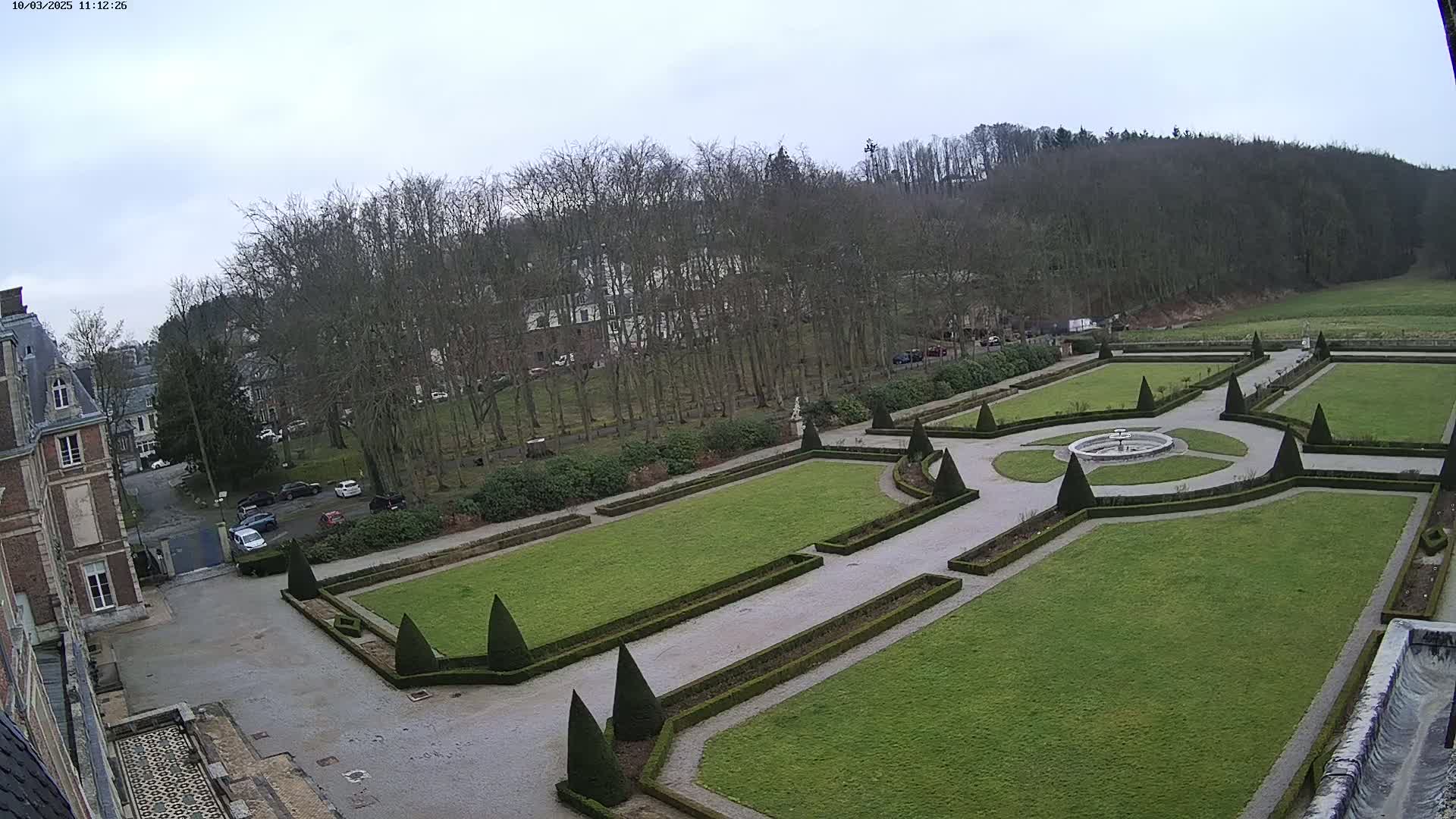 An overcast day reveals a formal garden with manicured lawns, hedges, and a central fountain, situated next to a large brick building and a line of leafless trees.