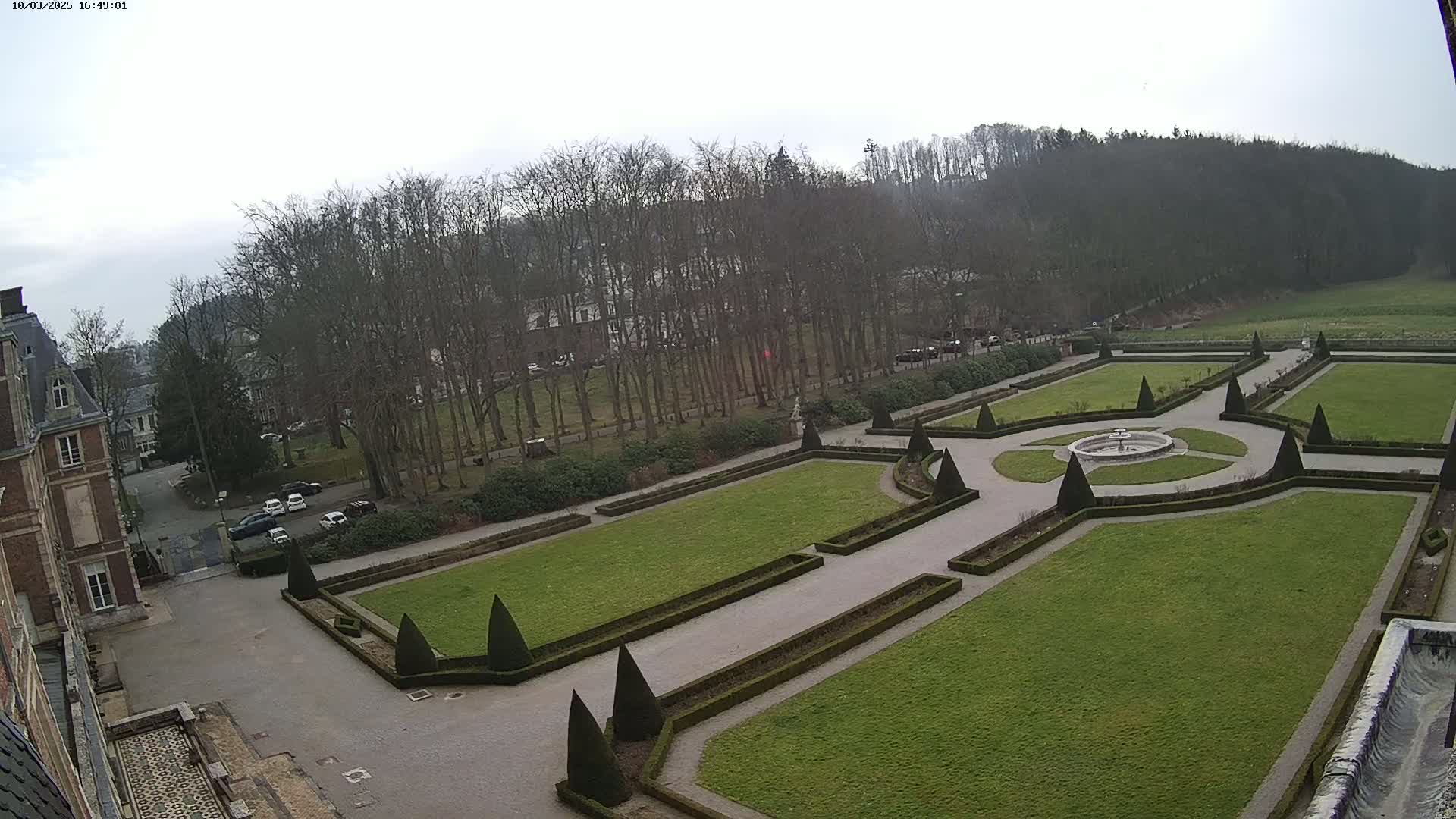 An overcast day reveals a formal garden with geometrically shaped lawns and hedges surrounding a central fountain, adjacent to a large brick building and a line of bare trees.