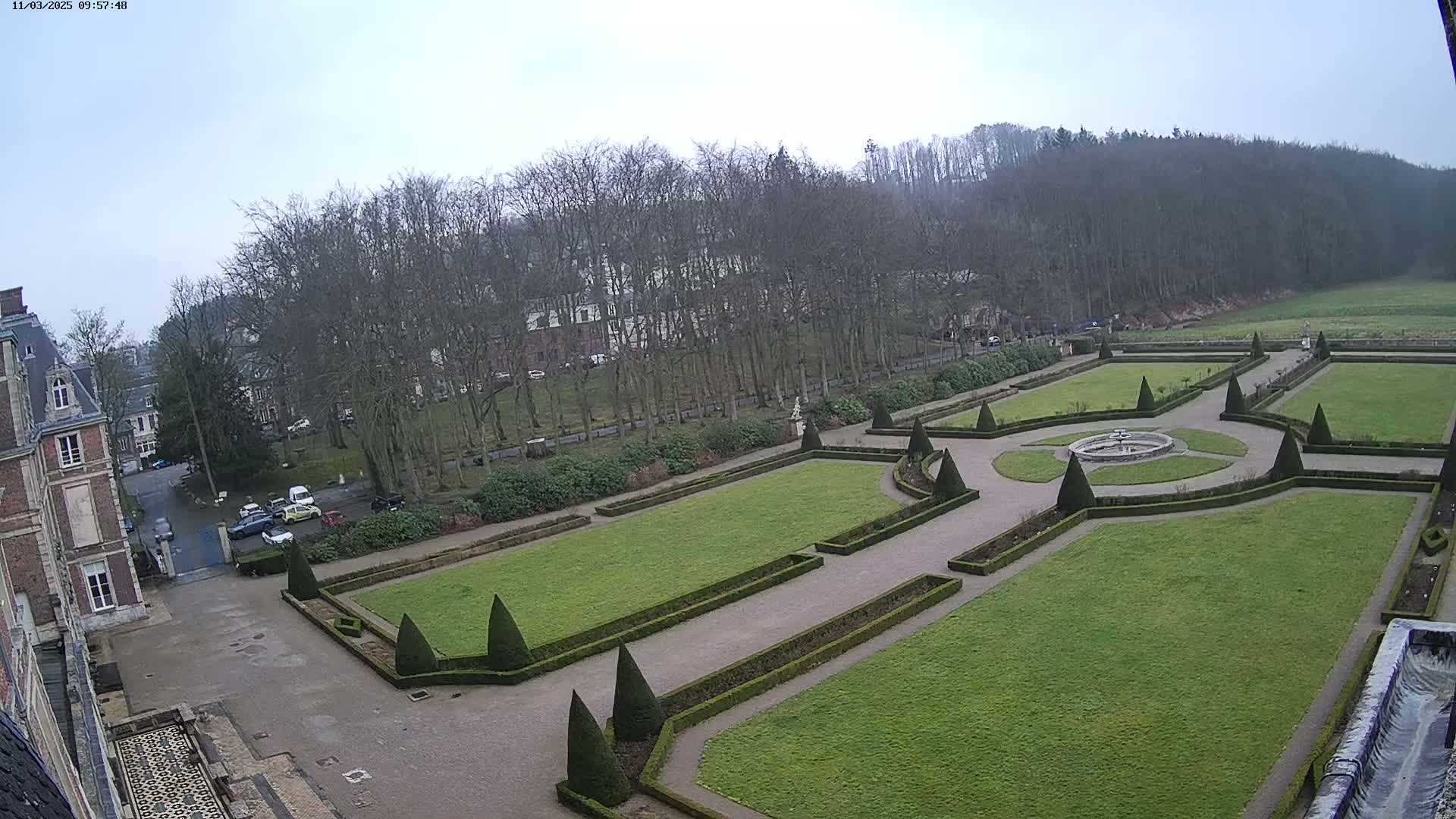 An overcast day reveals a meticulously manicured formal garden with geometric patterns of hedges and a central fountain, situated beside a large building and a line of trees leading to a hillside.