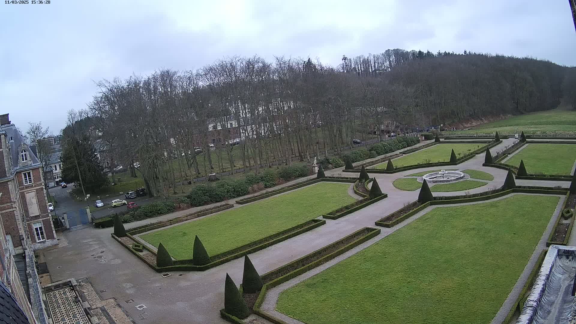 An overcast day reveals a formal garden with manicured lawns, hedges, and a central fountain, situated next to a large building and a wooded hillside.