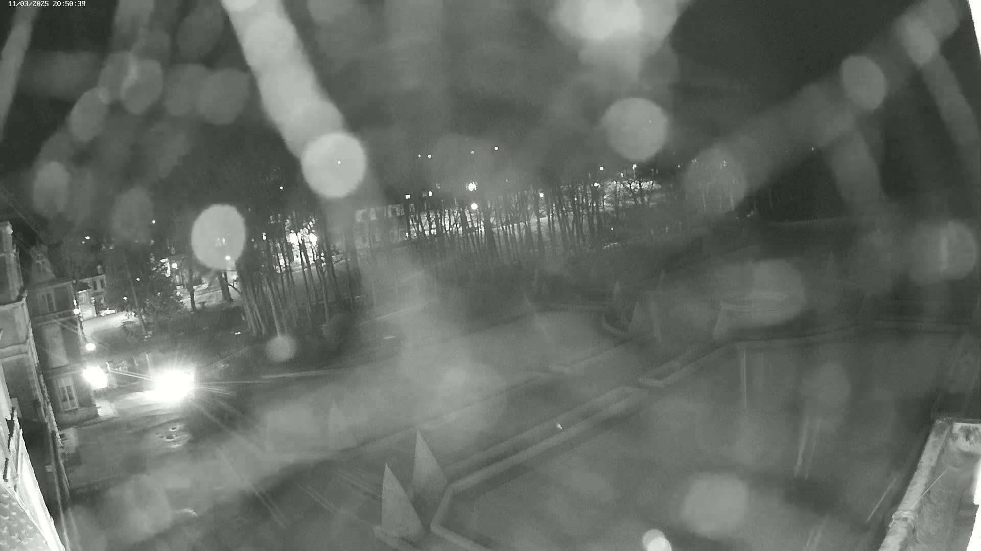 A nighttime, rainy view of a courtyard with geometric landscaping features and trees, partially obscured by rain on the camera lens.