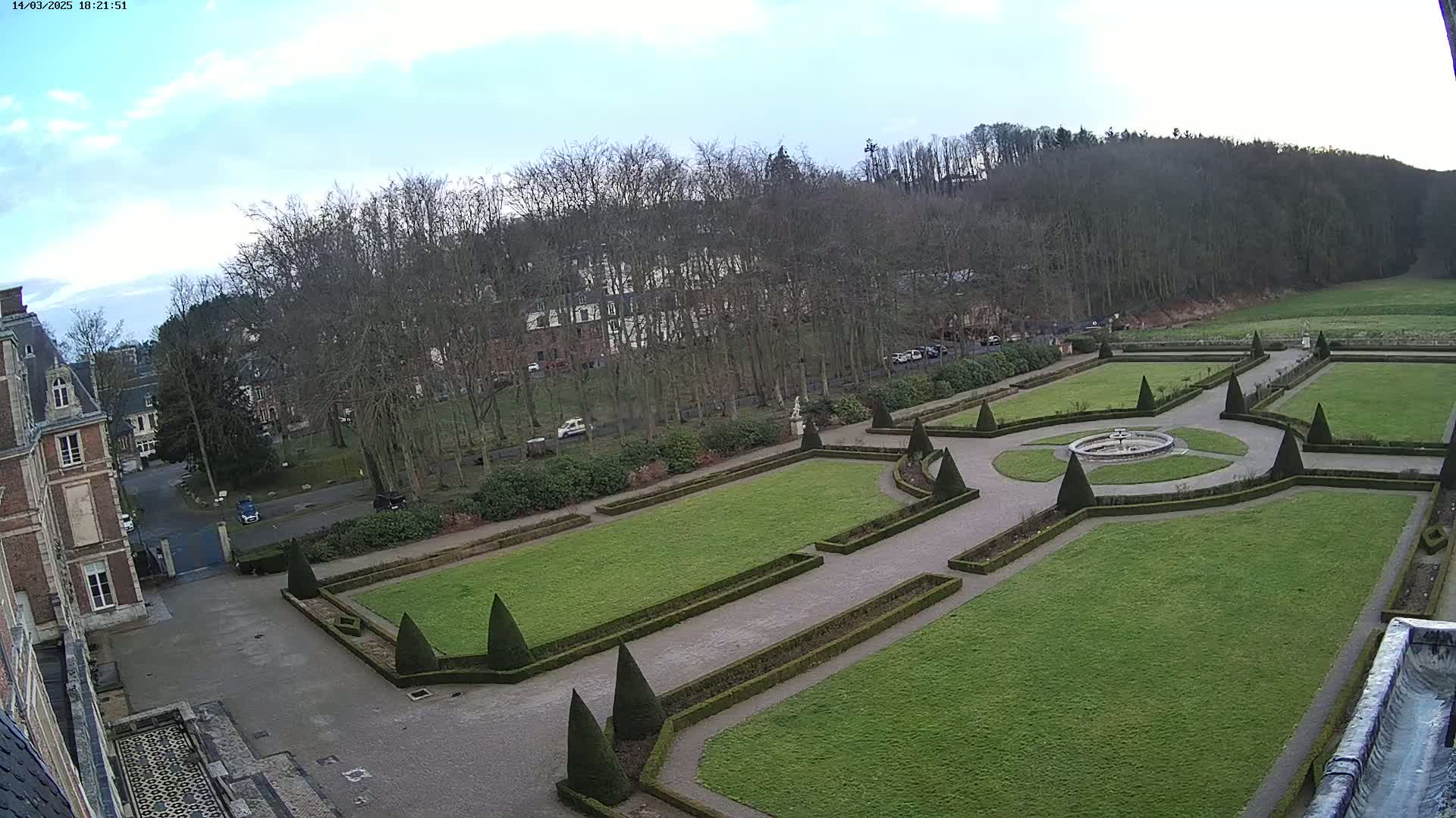 An aerial view shows a formal garden with manicured lawns, hedges, and a central fountain, adjacent to a large building and a wooded hillside under a partly cloudy sky.
