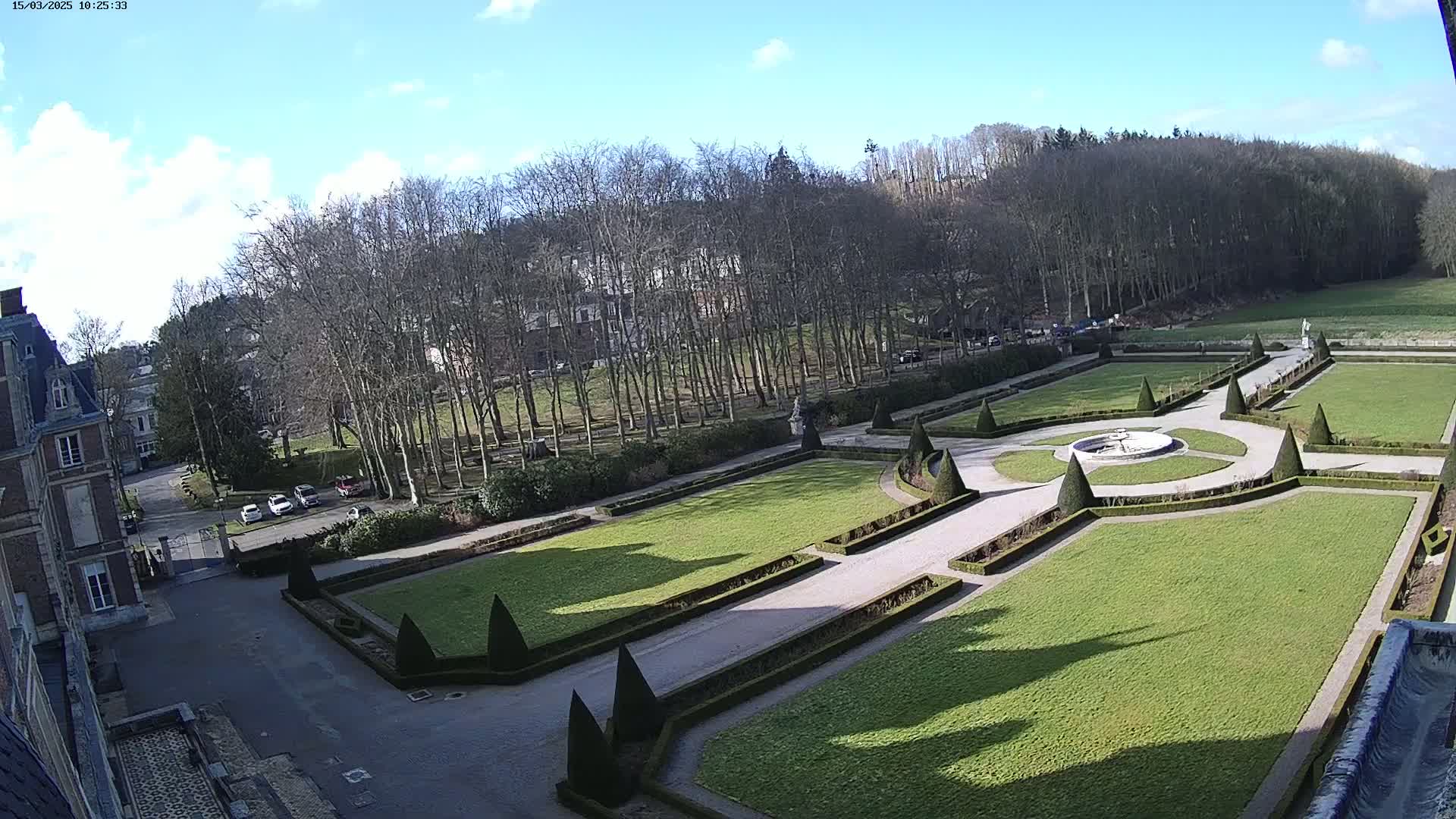 A formal garden with manicured lawns, hedges, and a fountain is visible from a high vantage point on a sunny day, with leafless trees in the background.