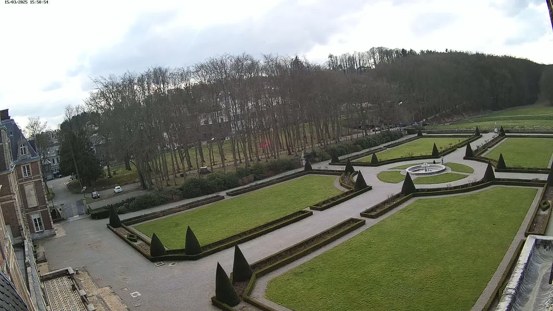 An aerial view of a formal garden with manicured lawns, hedges, and a central fountain, situated next to a building and a wooded area under an overcast sky.