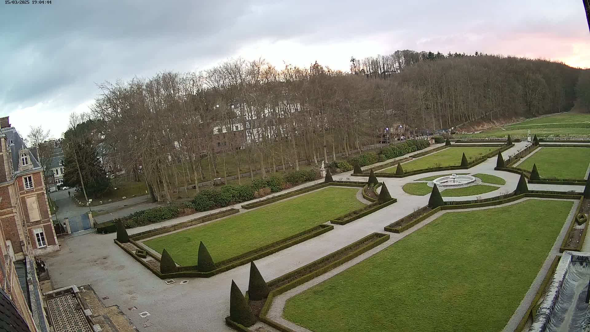 An aerial view of an ornate formal garden with manicured lawns, hedges, and a central fountain, set against a backdrop of bare trees under a cloudy sky.